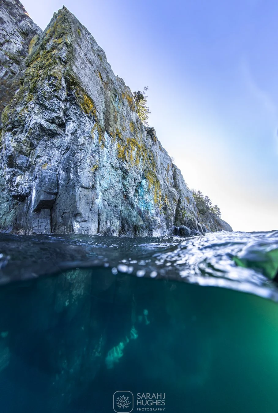 A rugged rocky cliff on Quadra Island rises from the water, with some greenery and trees on top. The photo is taken at water level, showing the boundary between water and rock, with a clear blue sky above.