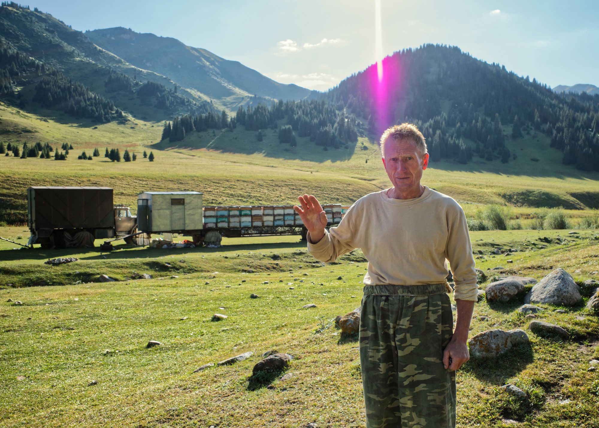 An elderly man in camouflage pants and a beige sweatshirt standing outdoors in a grassy field with mountains in the background. He is waving with his right hand and has a slightly puzzled or concerned expression. There are large rocks on the ground a