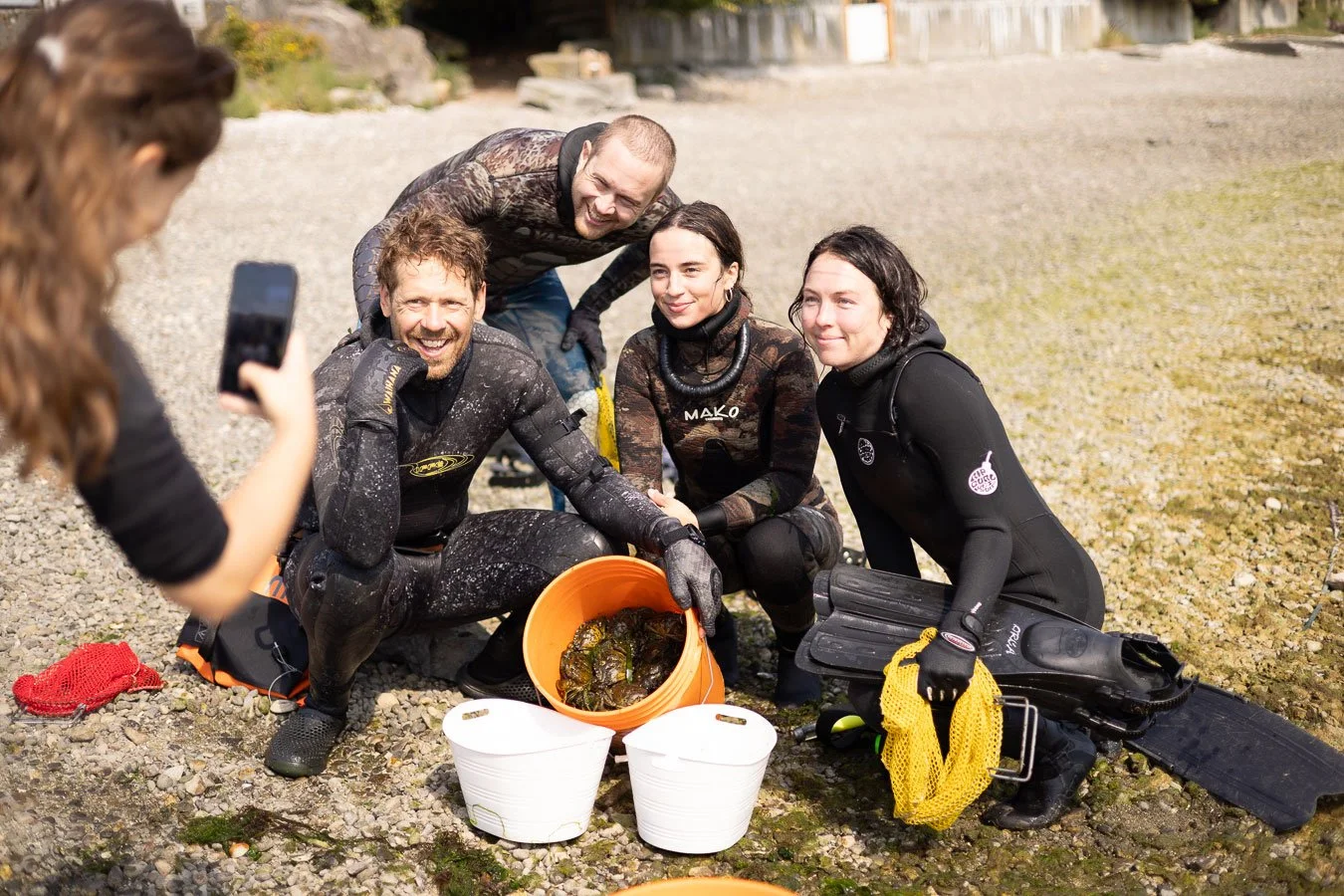 Group of people in wetsuits smiling and posing for a photo on a rocky beach with green crabs in buckets.