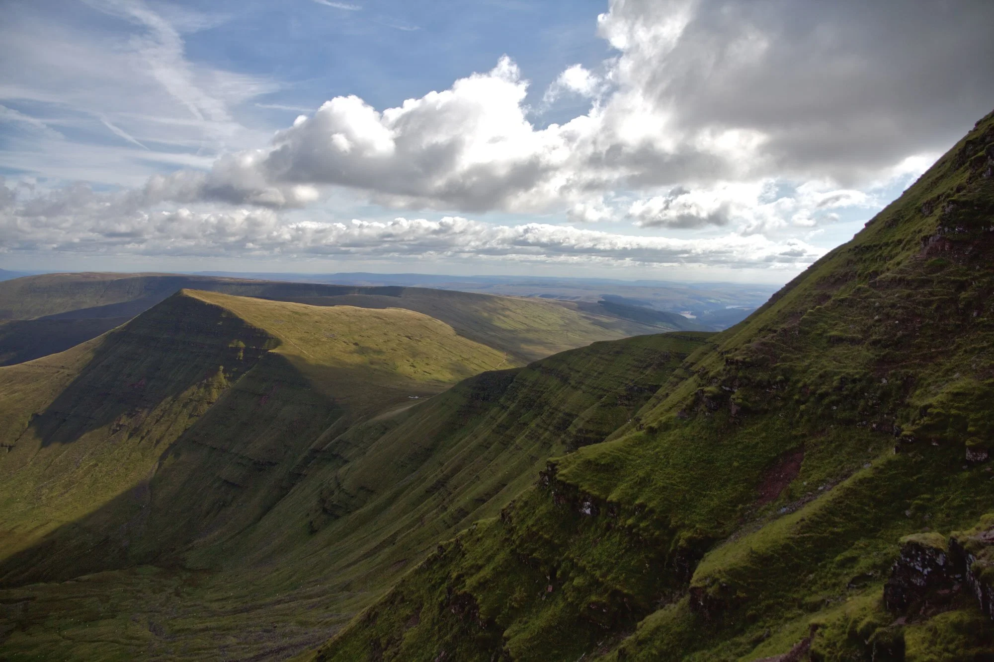 Green moss-covered mountainous landscape under a cloudy sky with patches of sunlight.