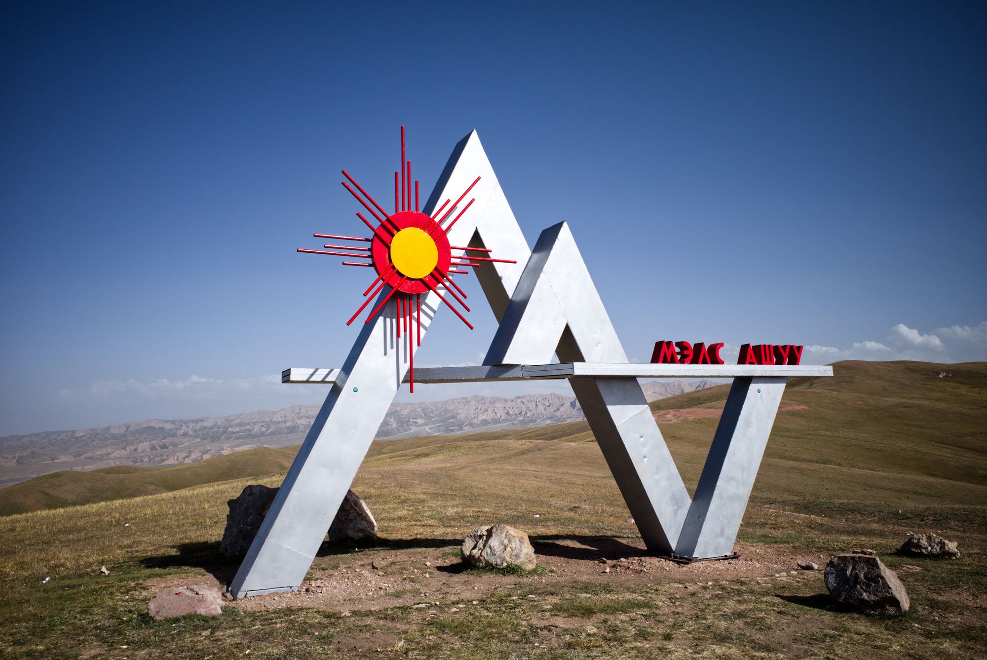 A modern outdoor sculpture with white angular supports, a red and yellow sunburst feature, and red letters spelling "MERCURY" in a green grassy landscape with mountains in the distance under a blue sky.