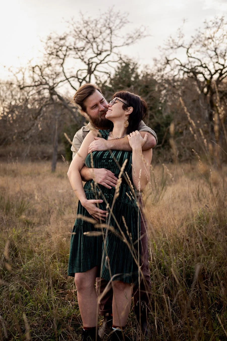 A couple embracing outdoors in a field with dry grass and leafless trees in the background at sunset.