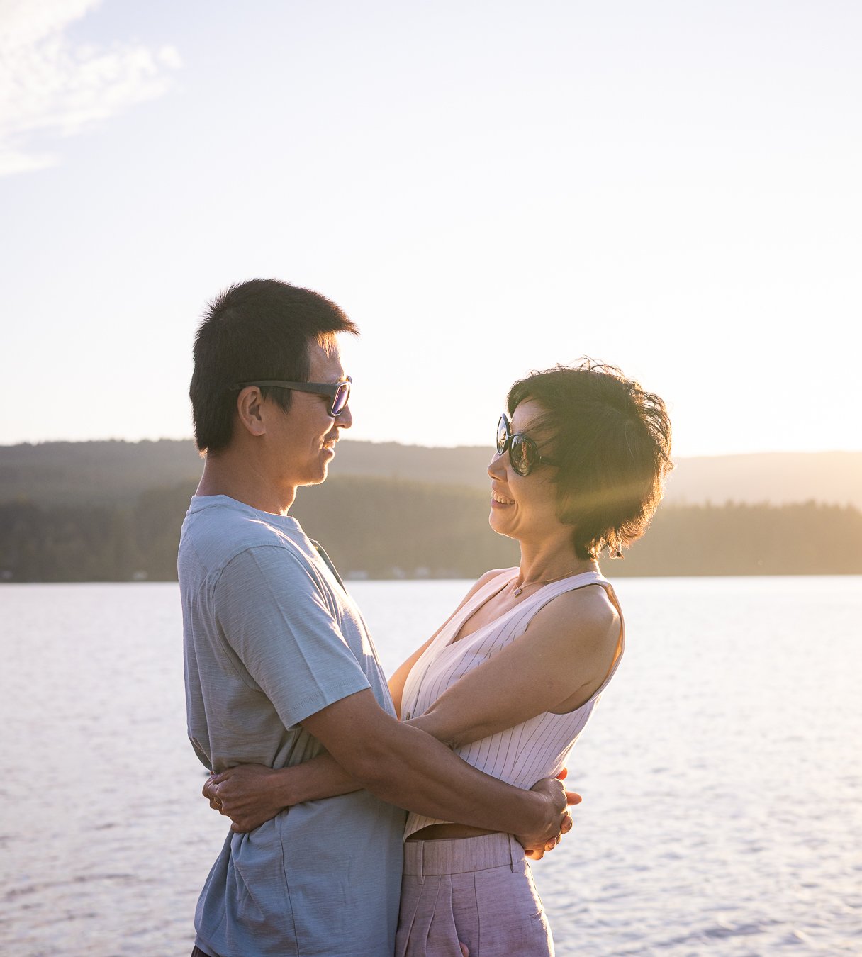 A middle-aged woman and a young man are standing close to each other near a body of water, smiling at each other during sunset, with the sun creating a warm glow in the background.