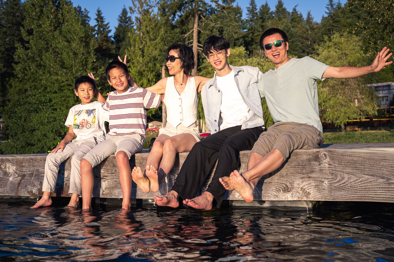 Family sitting on a dock with feet in the water, smiling and enjoying a sunny outdoor day.