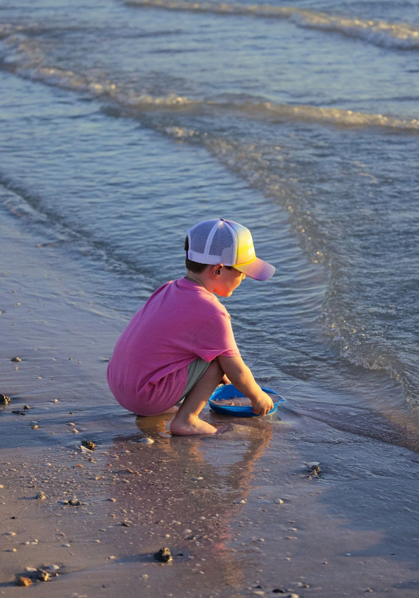 A young boy in a pink shirt and cap squatting on the beach, playing with a blue frisbee near the shoreline.