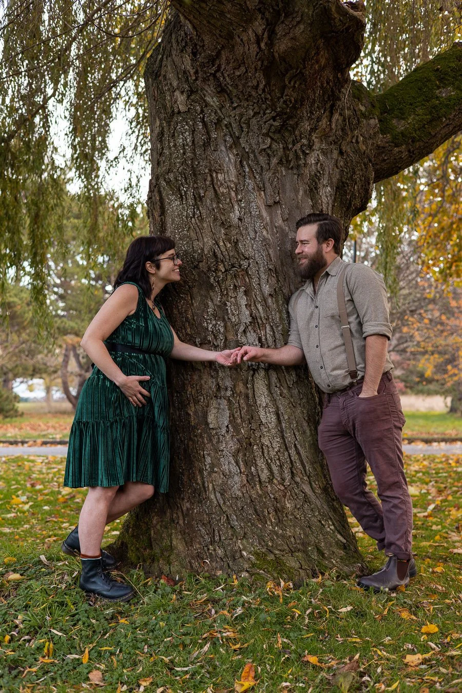 A man and woman standing next to a large tree in a park, holding hands and smiling at each other during fall.
