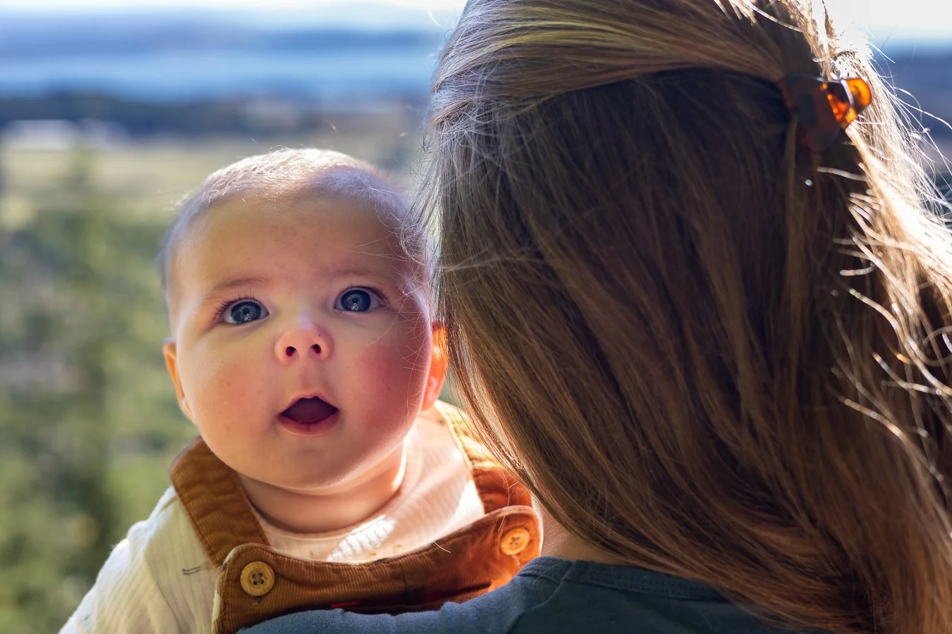 A baby with blue eyes and fair skin looking surprised, being held close to a woman with brown hair, outside on a sunny day.