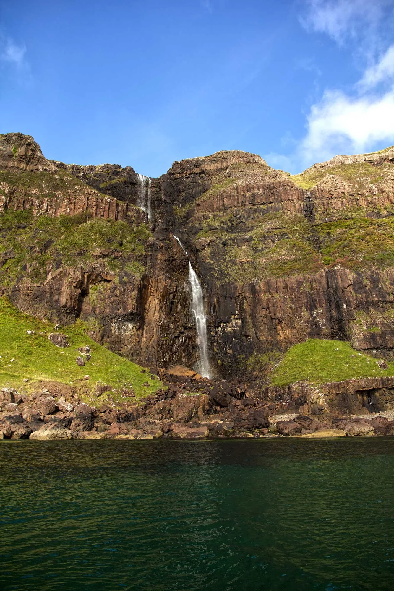 A waterfall flowing down a rocky, green hillside into a body of water, with a bright blue sky overhead.