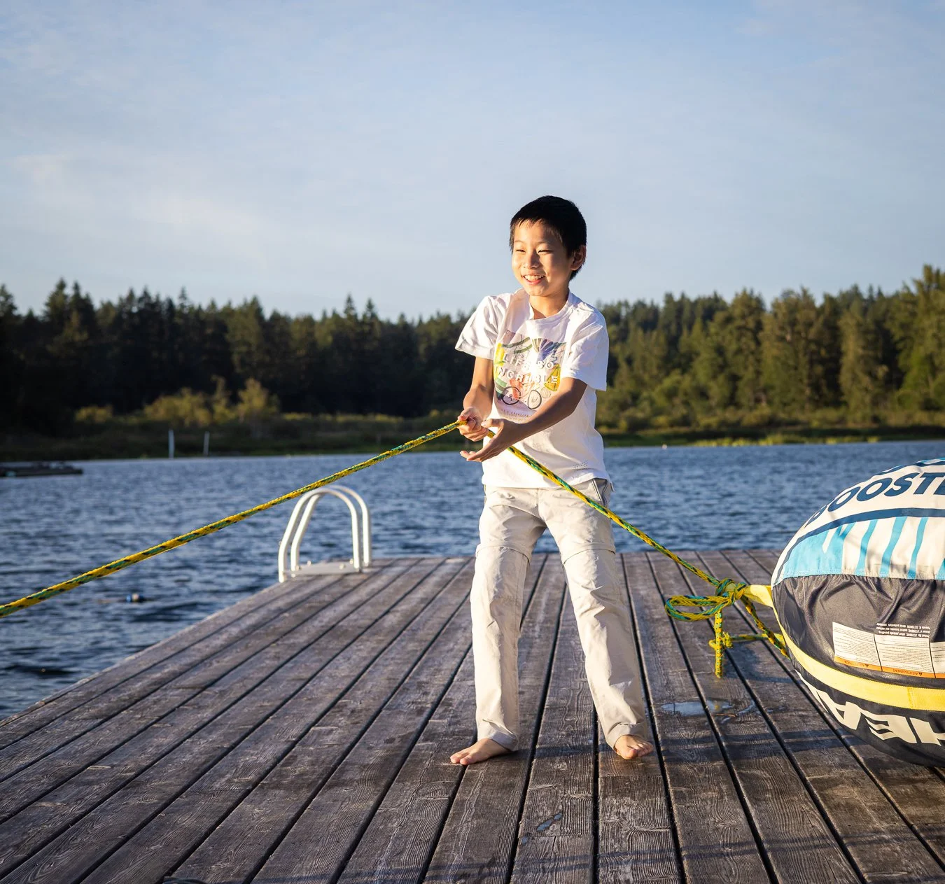 A young boy is smiling and pulling on a rope on a wooden dock by the water, with a bag nearby and trees in the background.
