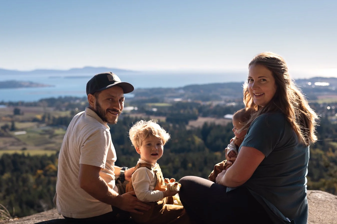 A family of four enjoying an outdoor view on a hillside, with a lake and mountains in the background. The father, mother, and two children are smiling and sitting on a rock.
