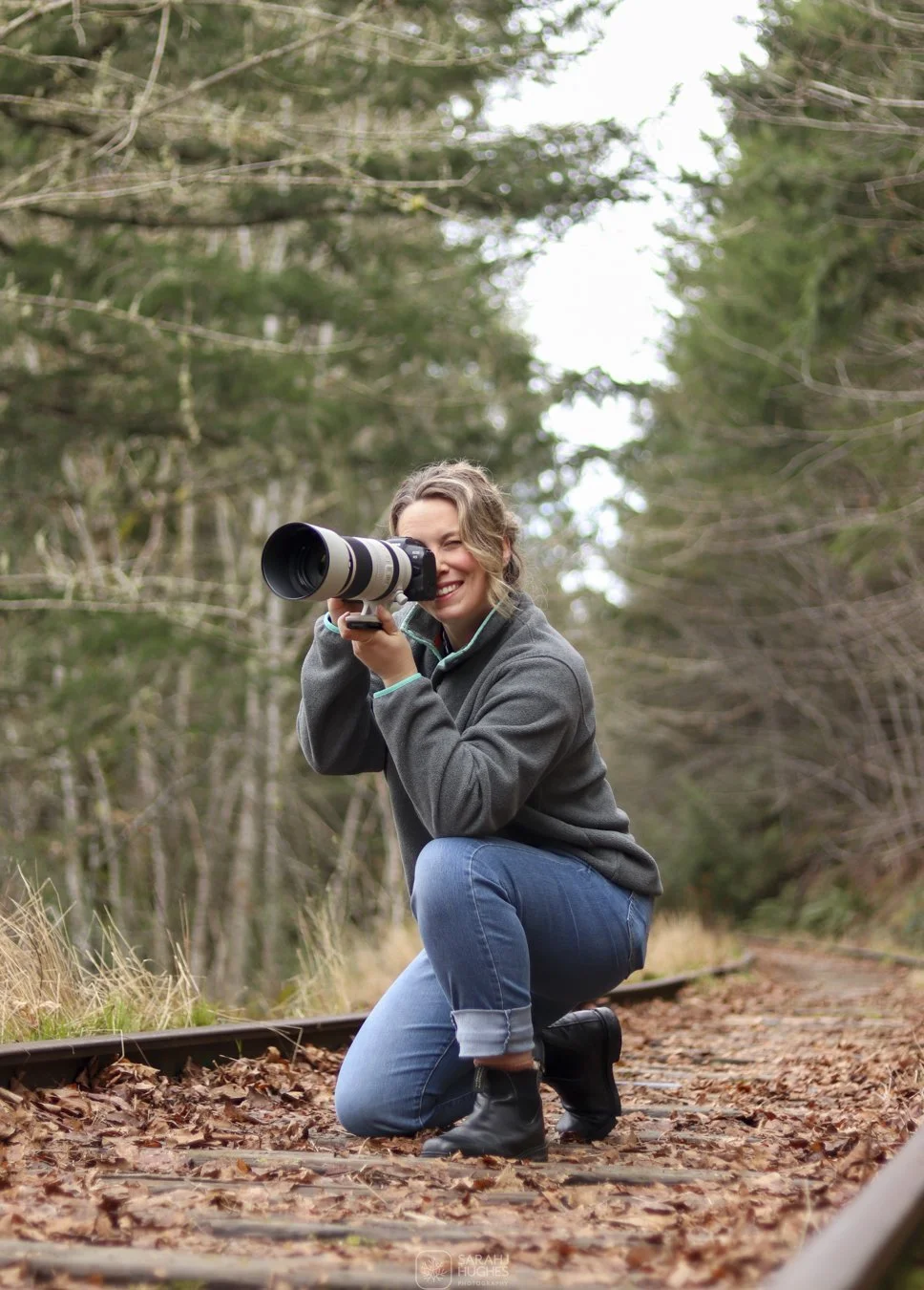 A woman, Sarah J Hughes, with blonde curly hair wearing a gray jacket, blue jeans, and black boots is kneeling on a railroad track in a forested area, taking a photograph with a large camera lens, smiling.