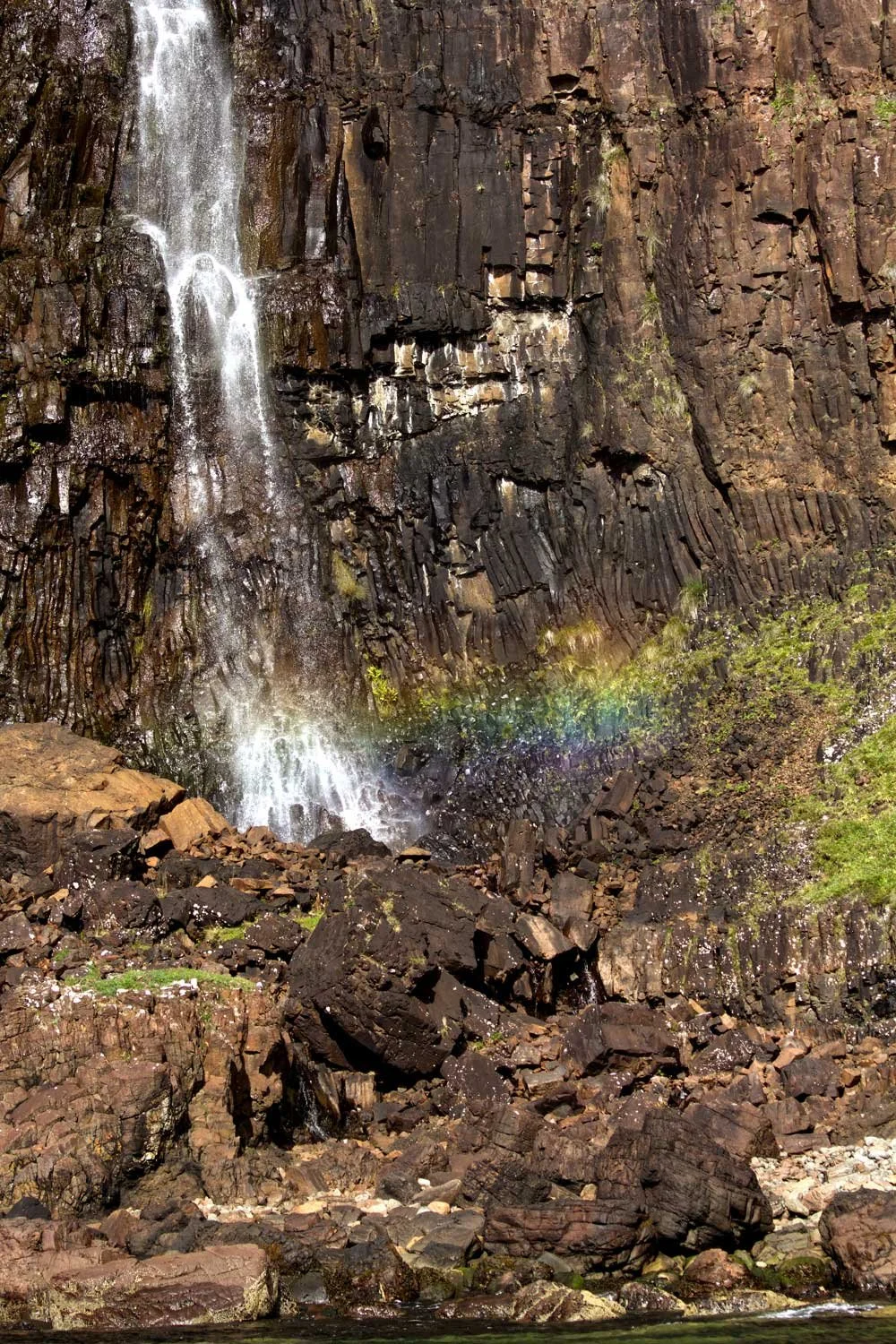 A waterfall cascading down a dark, rocky cliff with a rainbow near the bottom of the falls.