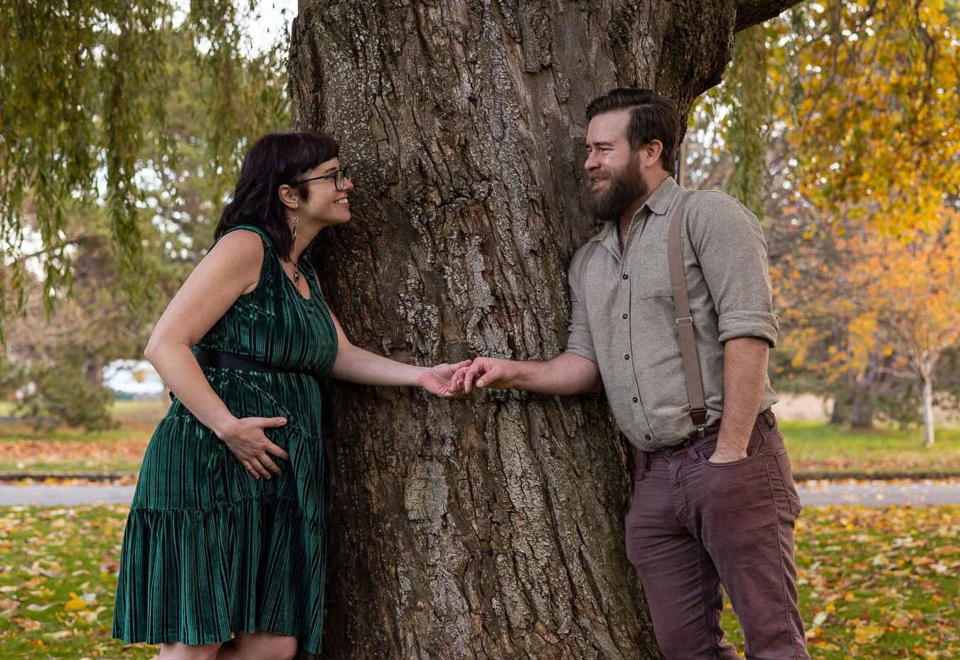 A woman in a green dress and glasses holding hands with a man with a beard, both smiling, standing on opposite sides of a large tree in a park with autumn foliage.