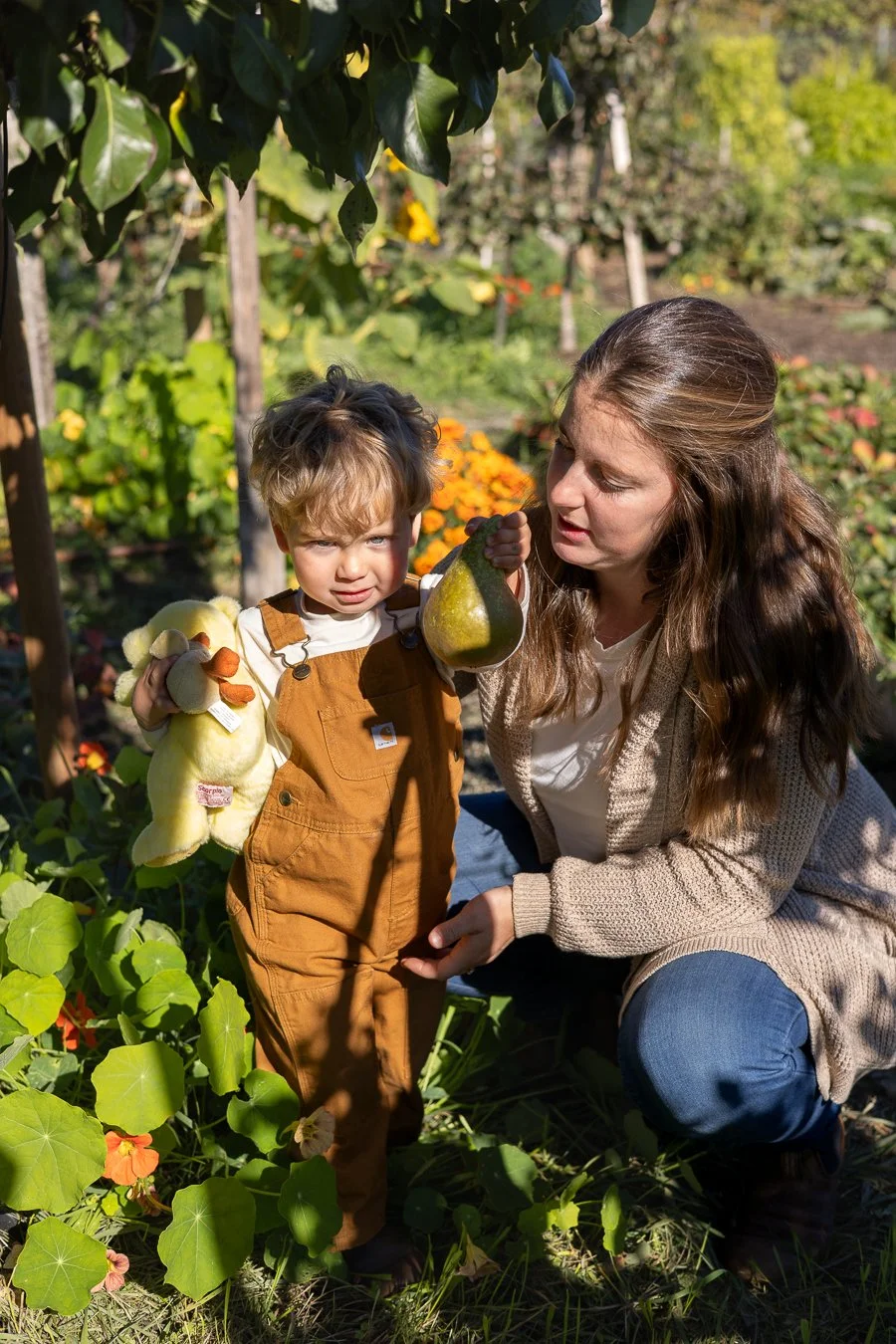 A woman and a young boy in a garden, with the woman holding a pear and the boy holding a stuffed animal, surrounded by green plants and flowers on a sunny day.