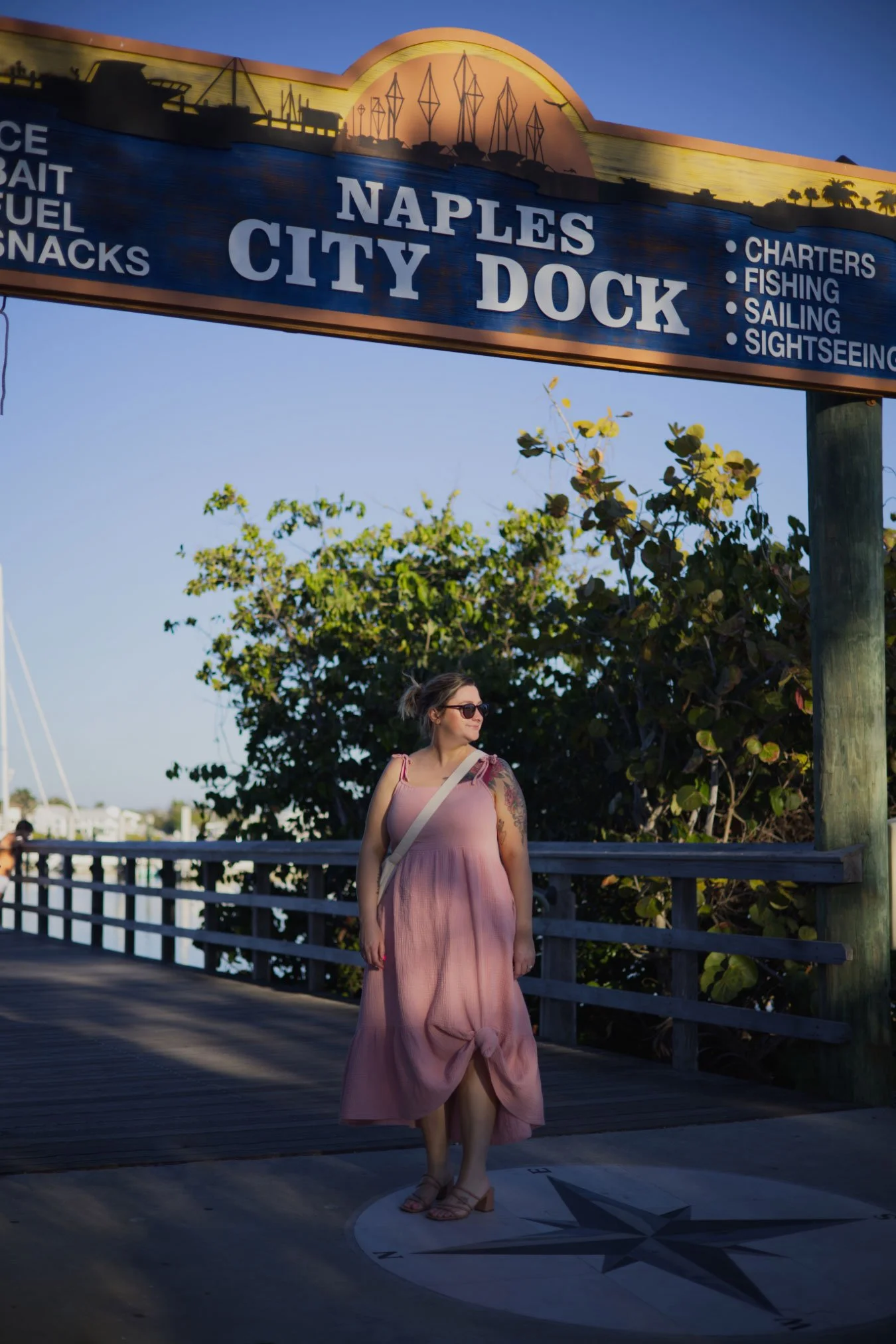 A woman in a pink sundress, wearing sunglasses and sandals, standing on a dock near greenery. The sign above reads 'Naples City Dock' with options for charters, fishing, sailing, and sightseeing.