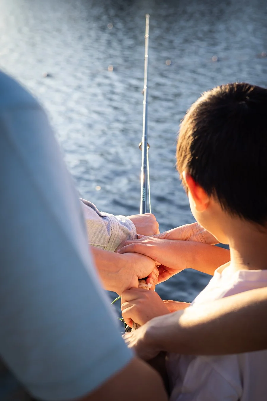 A young boy and an adult holding a fishing rod and a fish near the water.
