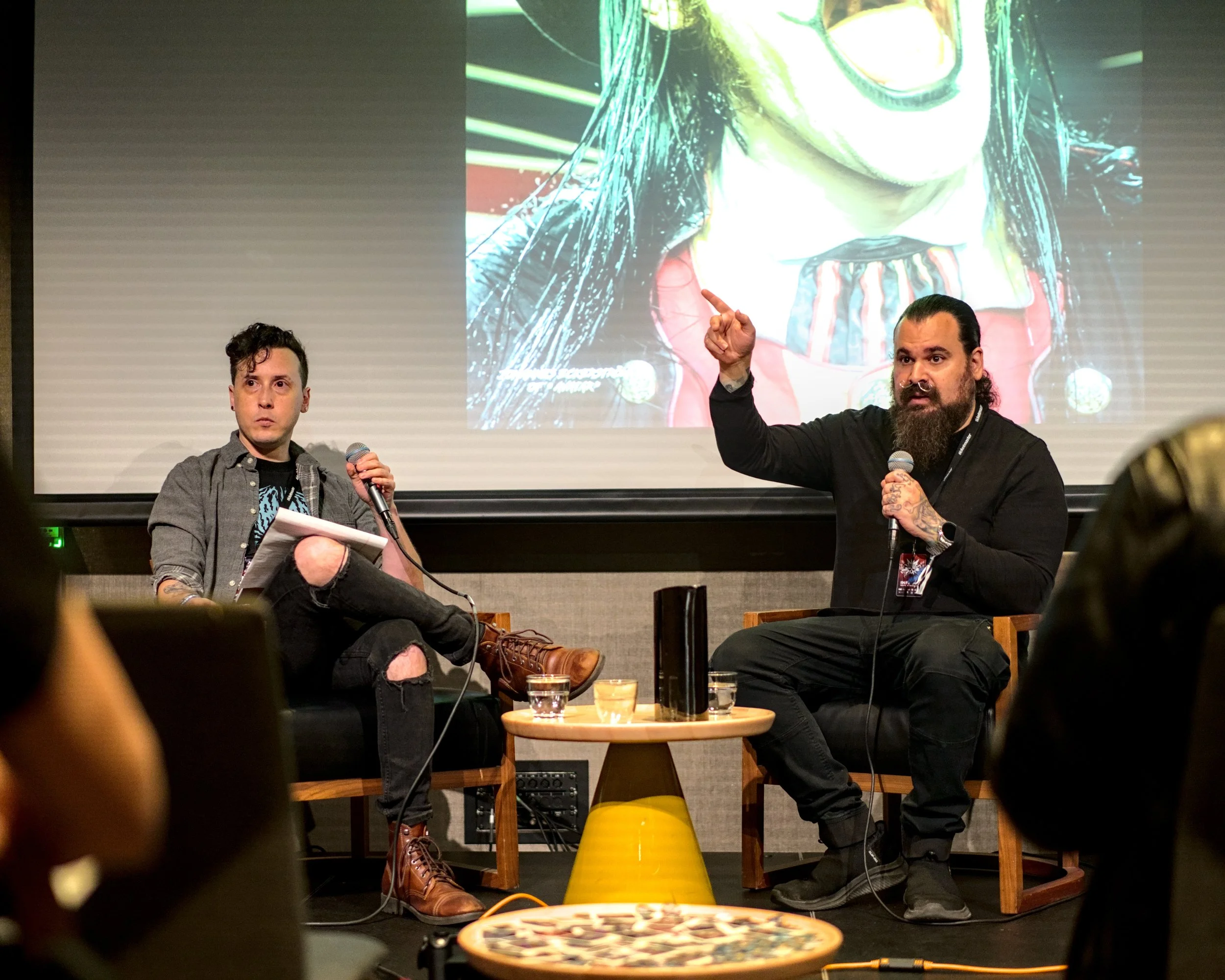 Two men sitting on a stage in front of a large screen, engaged in a discussion. The man on the right is speaking into a microphone and gesturing with his hand. The man on the left is holding a microphone and a notepad, looking attentive. The background features an animated image of a clown on the screen.