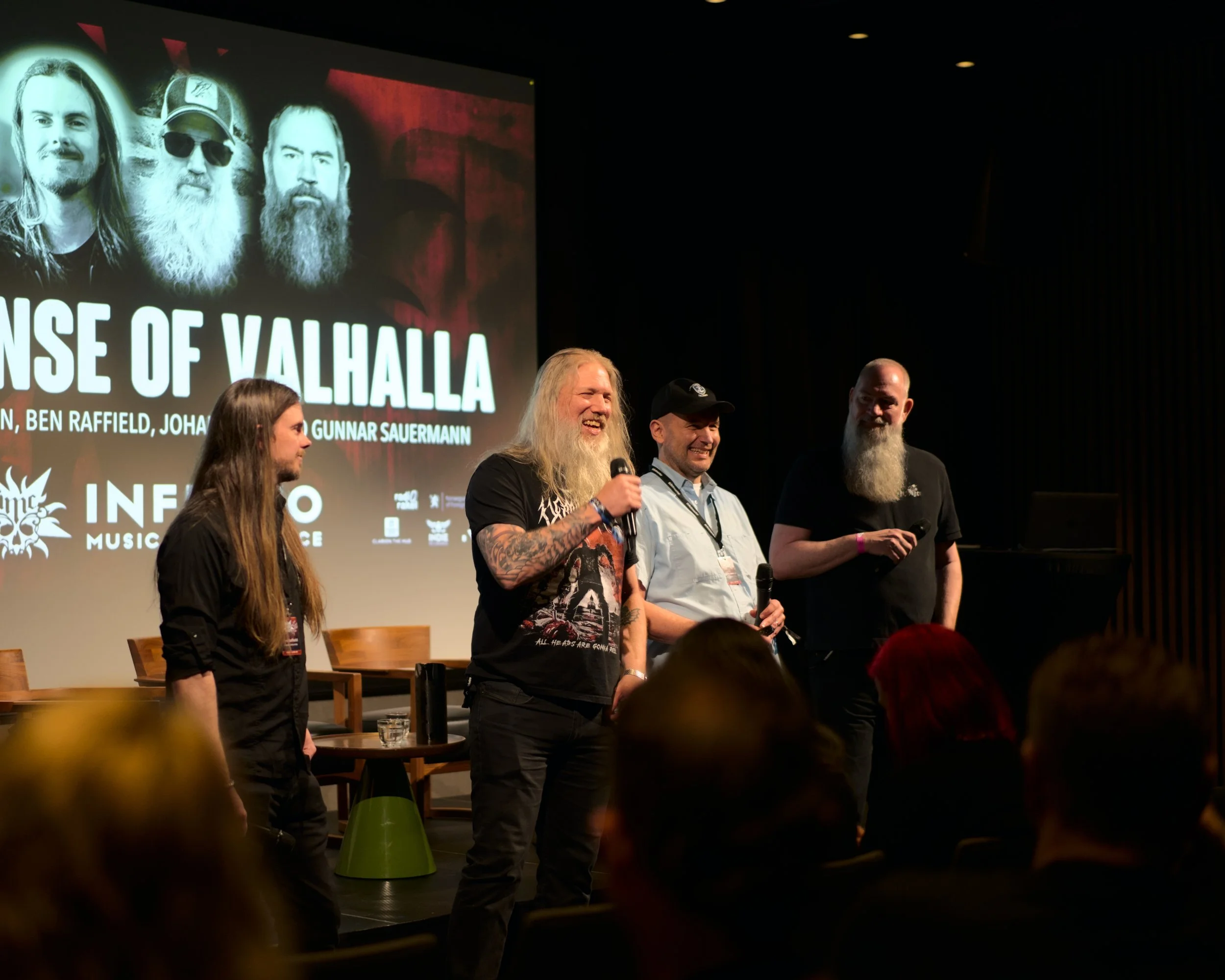 Four people standing on stage at a panel discussion, with a large screen behind them displaying the title 'RANS OF VALHALLA' and images of four men with long hair and beards.