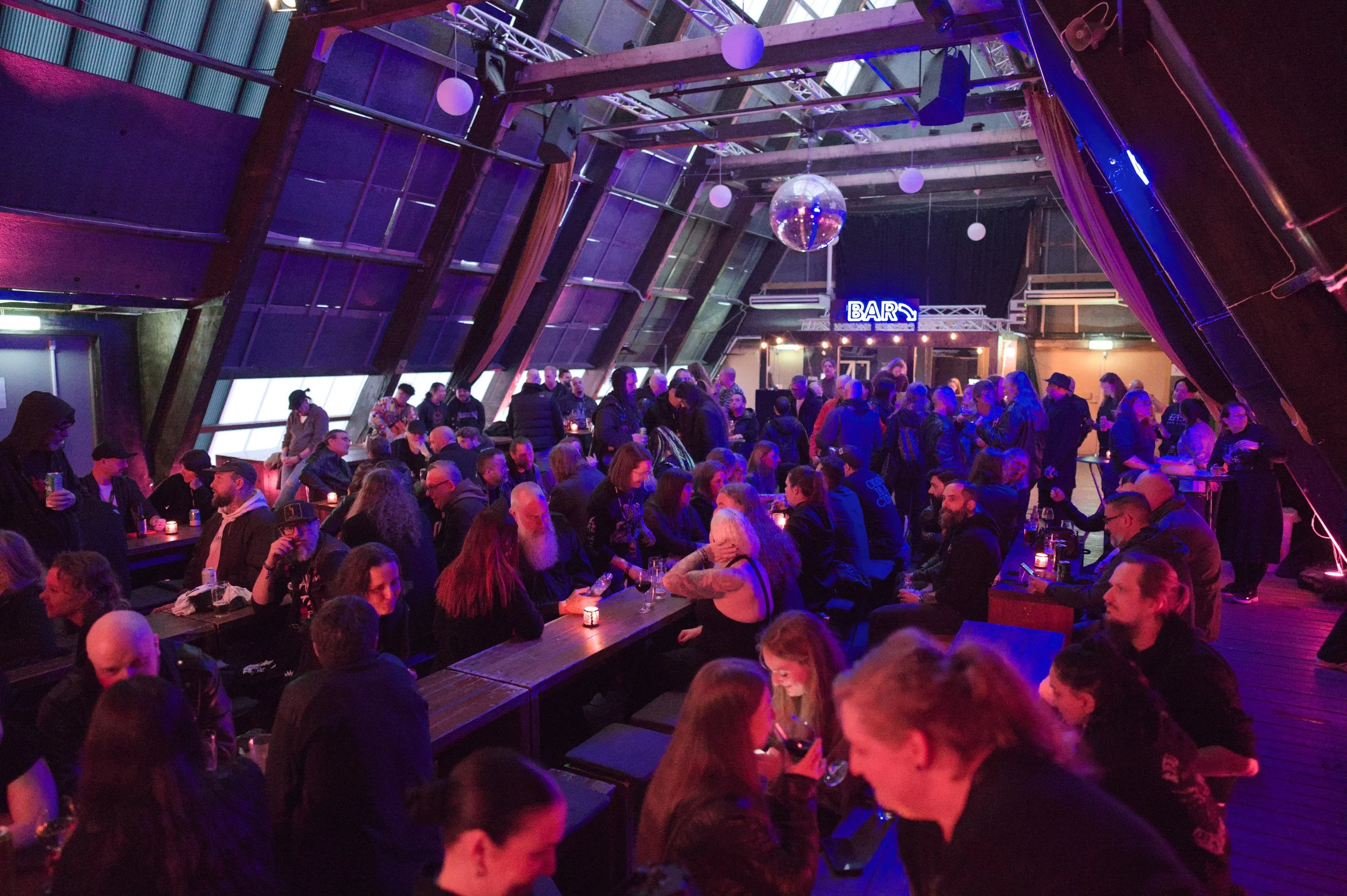 Crowded nightclub with people sitting at tables and standing, dim lighting, purple and blue hues, a large disco ball hanging from the ceiling, and a neon sign that says 'BAR' in the background.