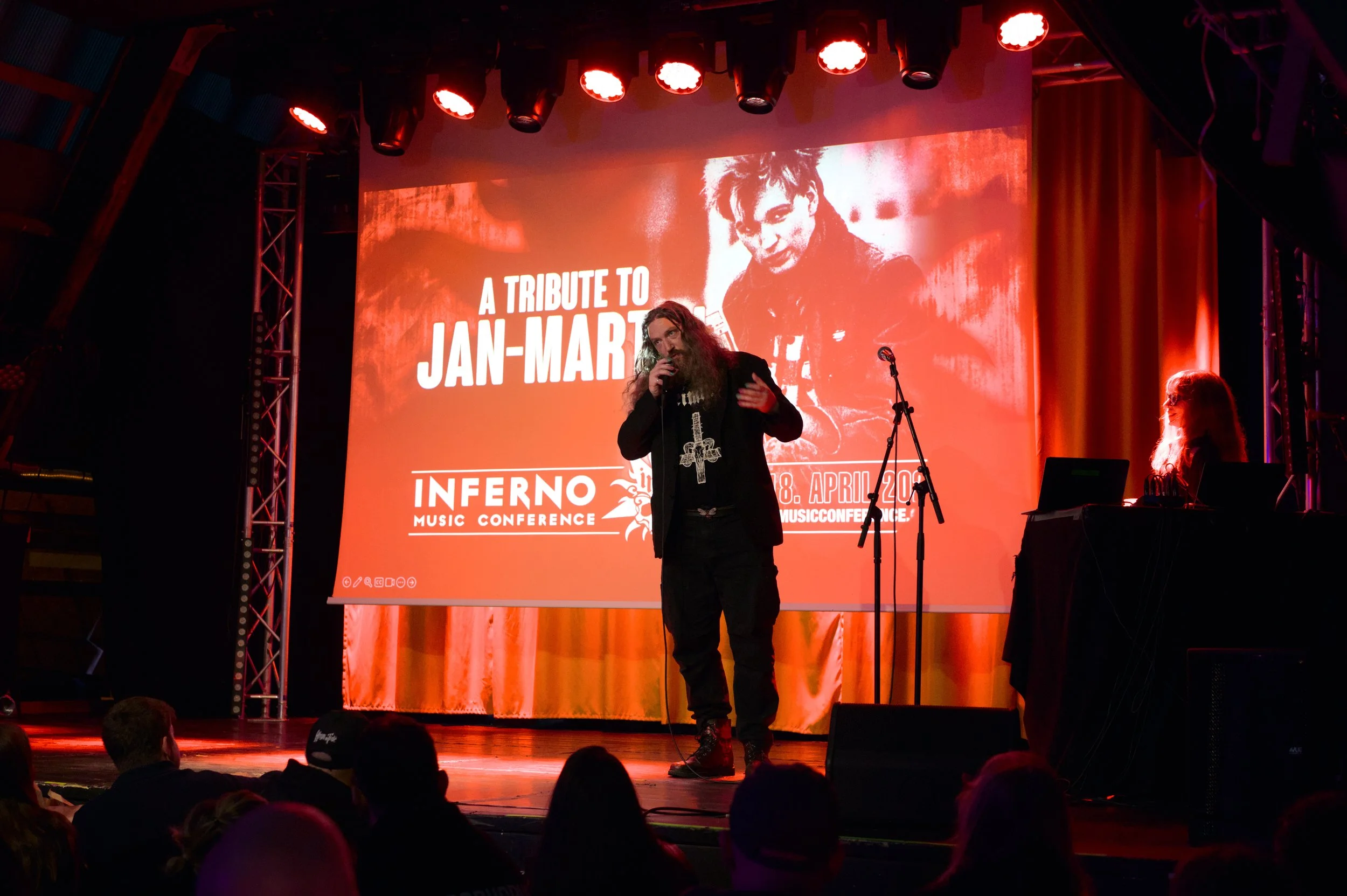A man with long hair and a beard speaking into a microphone on stage at the Inferno Music Conference, with a woman at a DJ booth nearby. The stage features a large screen with red background displaying text about a Jan-Marth tribute and the conference dates.