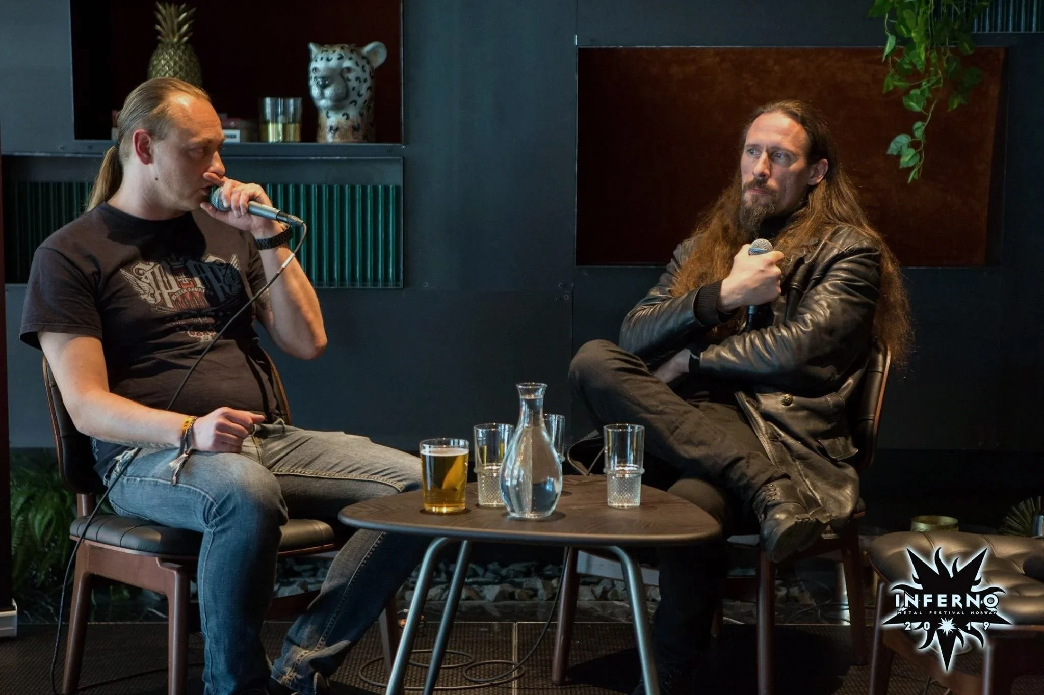 Two men sitting on chairs having a conversation with microphones, in a darkroom with black walls and decorative items. One man has long hair and is wearing a black leather jacket, the other has long hair tied back and is wearing a graphic T-shirt and jeans. There is a small logo at the bottom right corner reading 'Inferno Metal Festival Norway 2019'.
