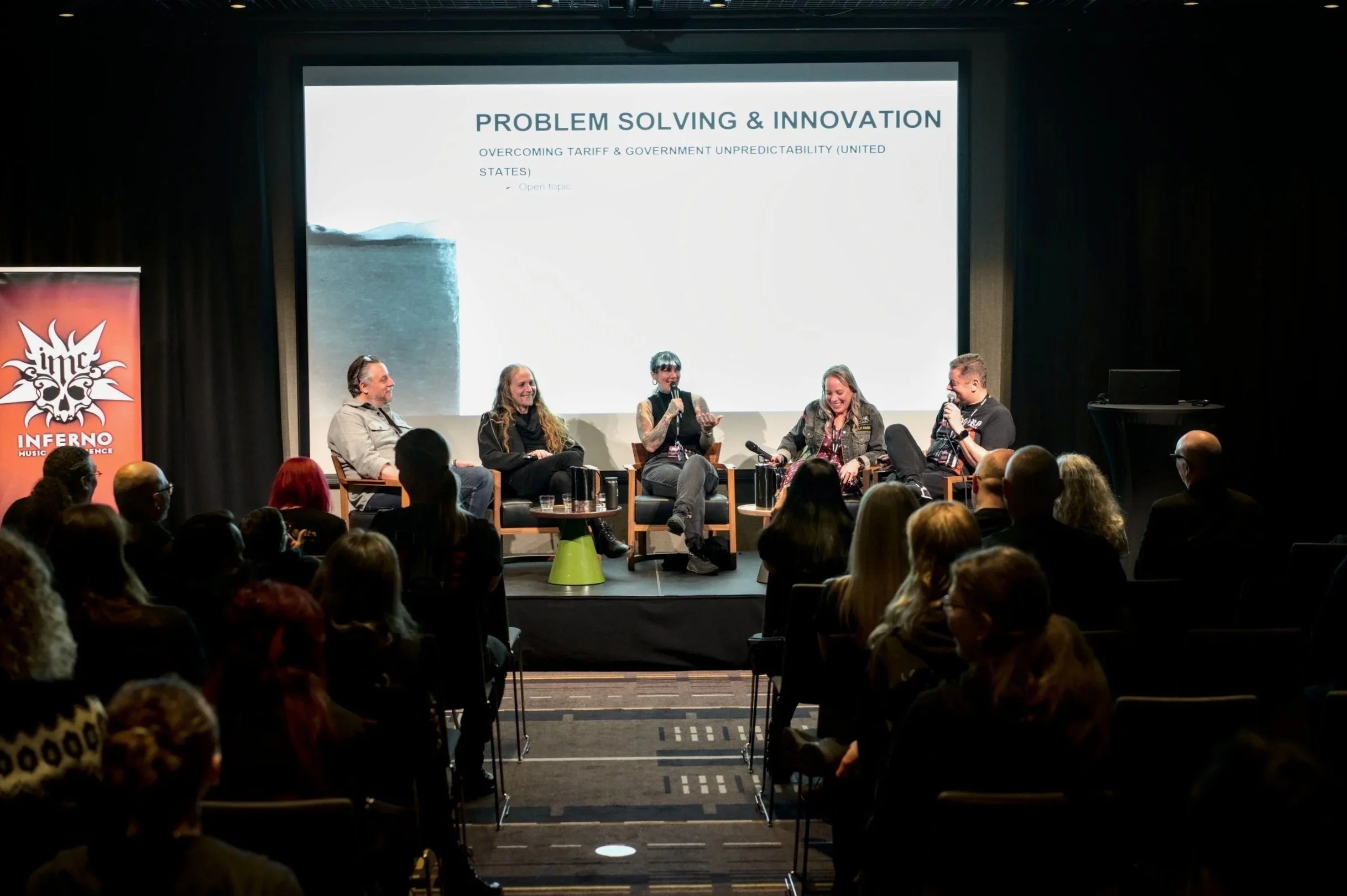 Panel discussion on stage at a conference with five women seated in armchairs, one woman speaking into a microphone, a large screen behind displaying a slide titled 'Problem Solving & Innovation,' audience in the foreground, and a red banner with a skull graphic and the word 'Inferno' to the left.