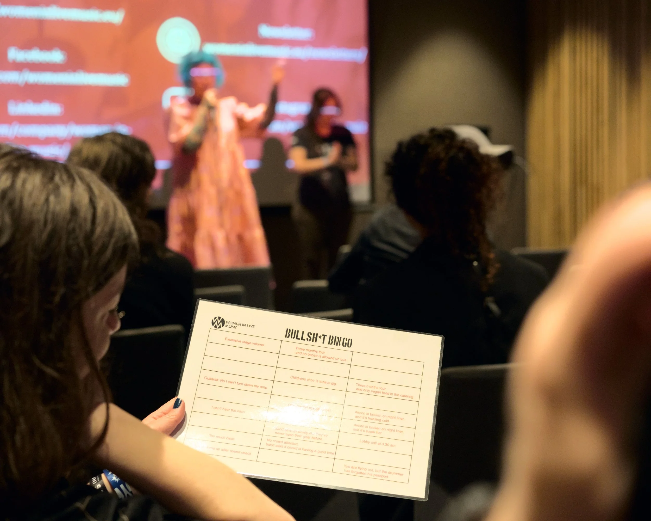 A woman in the audience holding a paper titled 'BULLSH*T BINGO' during a presentation or event, with a speaker on stage and a blurred screen in the background.