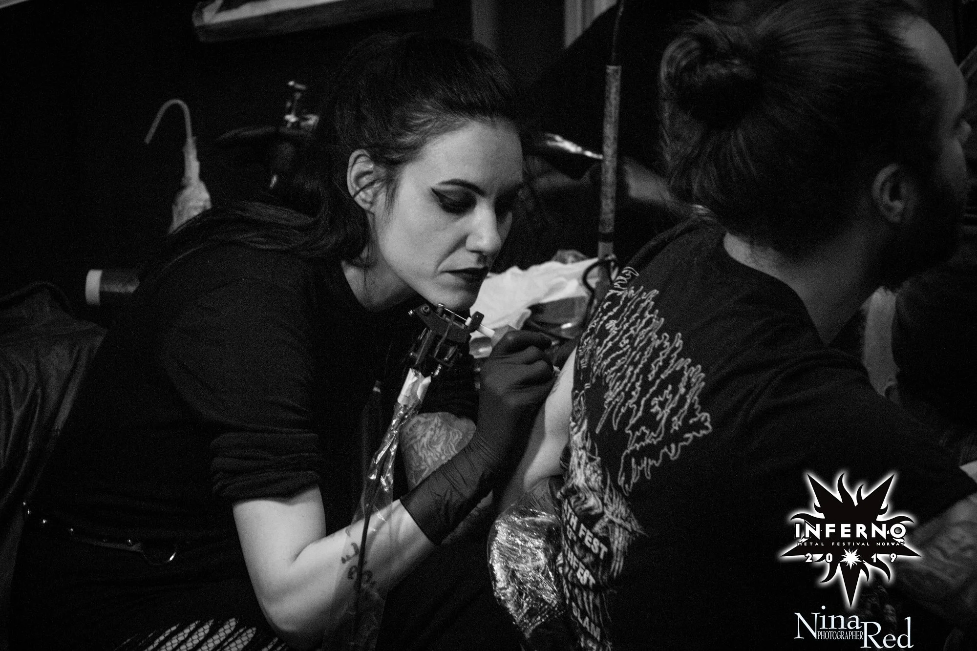 A woman with dark hair, makeup, and piercings is tattooing a person's arm at a metal festival.