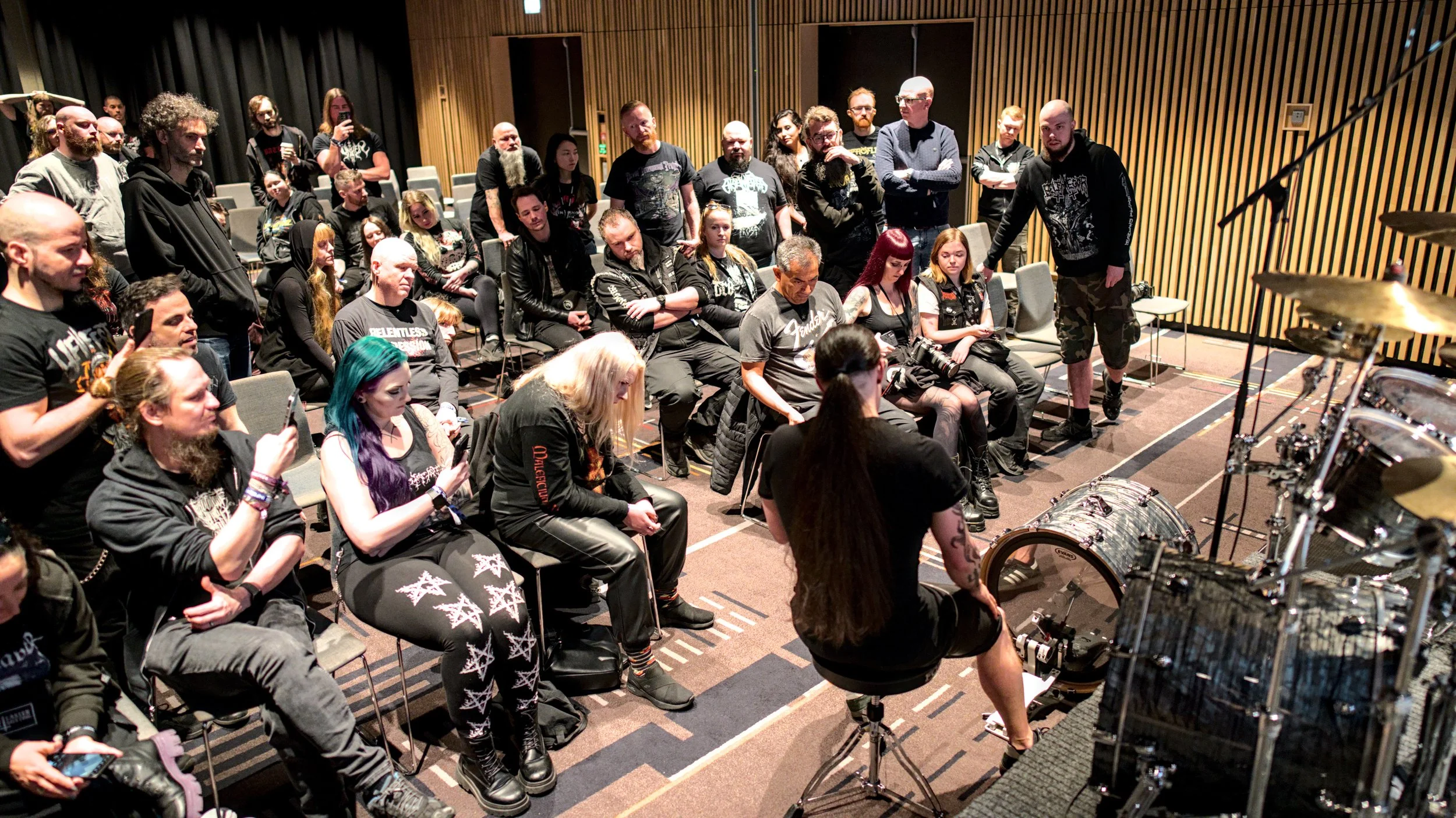 A group of people attending a music rehearsal or soundcheck in a room with wooden paneling walls. Some people are sitting, some standing, and they are focusing on a woman sitting near the drum set.