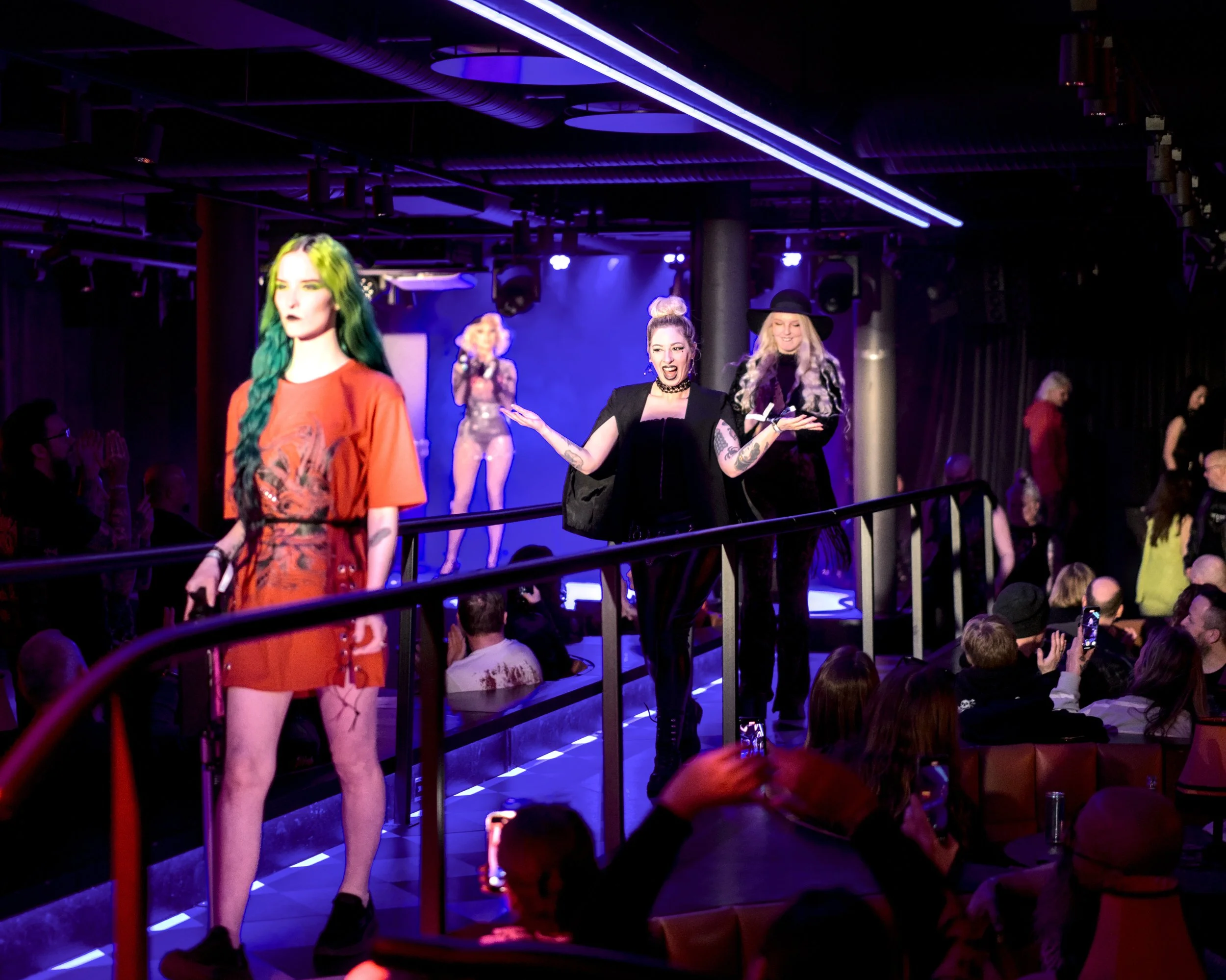 Performers on a stage during a fashion show with audience members taking photos.