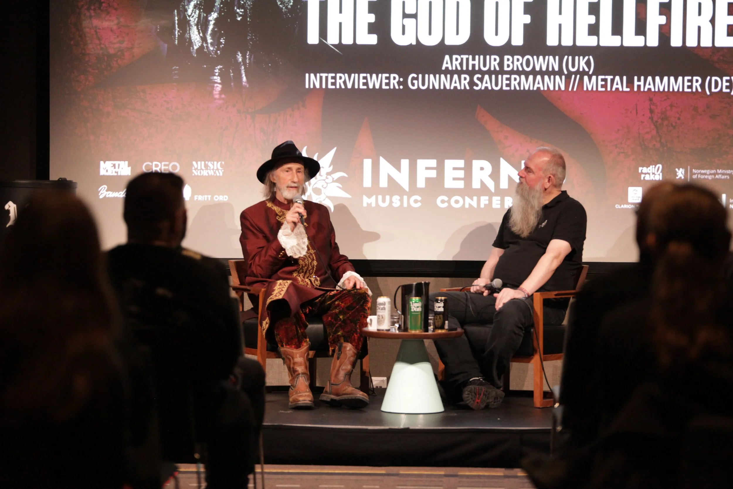 A man and a woman are seated on stage during an interview at the Inferno Music Conference. The woman is wearing a costume with a hat, patterned pants, and boots, and is speaking into a microphone. The man is dressed casually with a long gray beard and is listening. There is a large screen behind them displaying text about the event and participants.