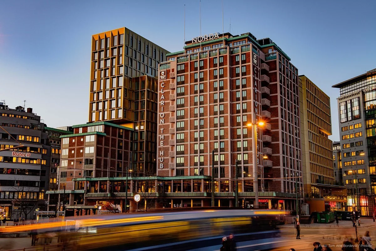 Cityscape of a downtown area at dusk featuring a tall brick building with vertical signage reading 'the Hub' and another building labeled 'Nordda'. Cars with streaks of light pass on the street, and illuminated street lamps and storefronts are visible.