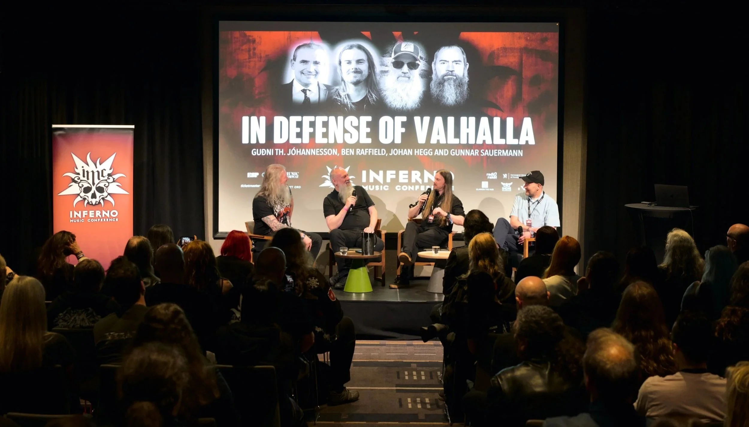 Panel discussion at a conference with four men on stage, two of whom are speaking into microphones. The background features a large screen with a promotional poster for a music conference titled "In Defense of Valhalla," and the banner on the left displays the conference logo and name.