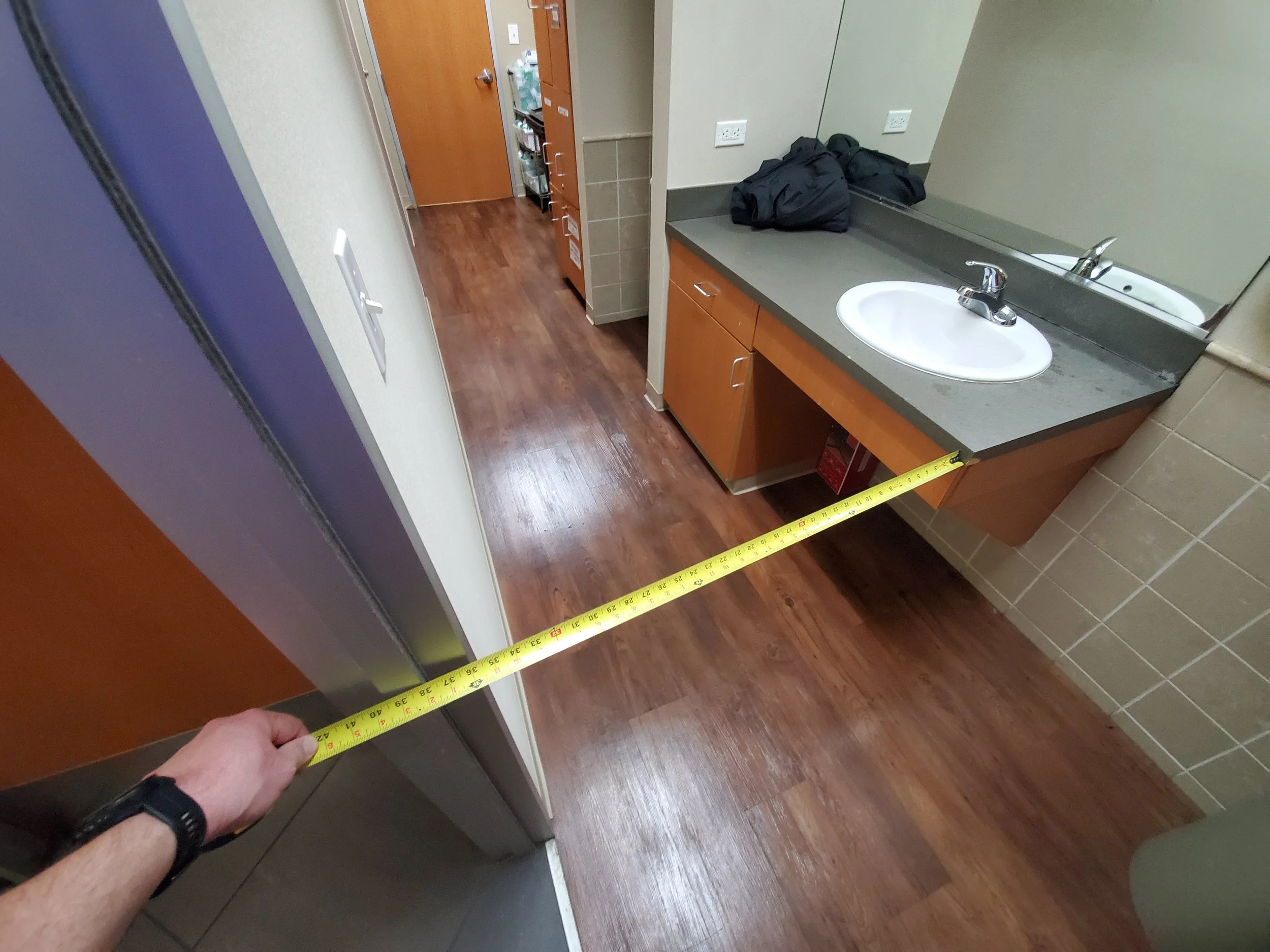 A person holding a measuring tape across a bathroom floor near a sink and mirror, measuring the space between the sink and the adjacent wall.