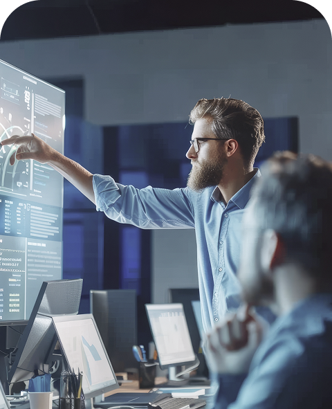 Man with glasses and beard pointing at a large digital screen displaying data and charts in a modern office with colleagues.