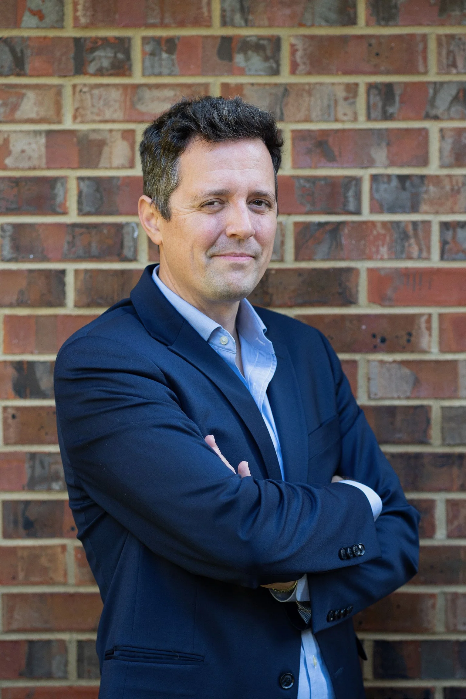 A man in a navy blazer and light blue shirt standing with arms crossed in front of a brick wall.