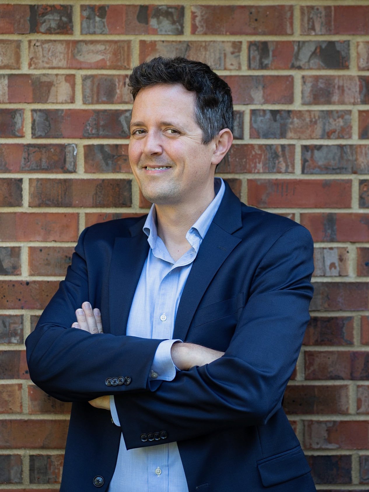 A man in a navy blazer and light blue shirt standing with arms crossed in front of a brick wall, smiling.