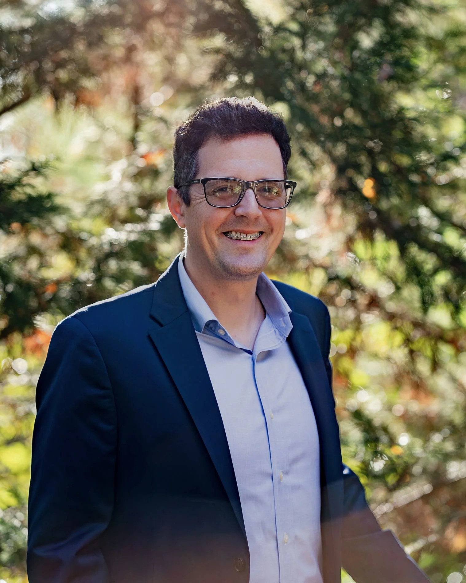 A smiling man wearing glasses, a navy blazer, and a light blue shirt outdoors among trees with sunlight filtering through the foliage.