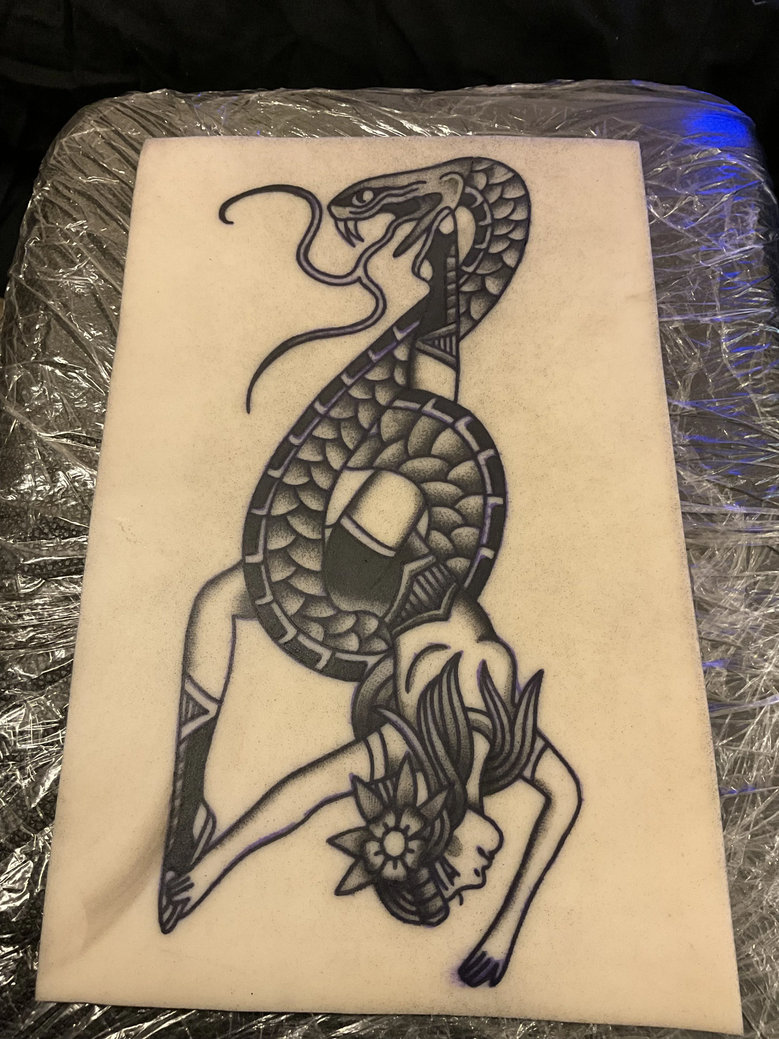 A tattoo design of a woman with long flowing hair and a flower in her hair, with a snake wrapping around her body and head, on a white background.