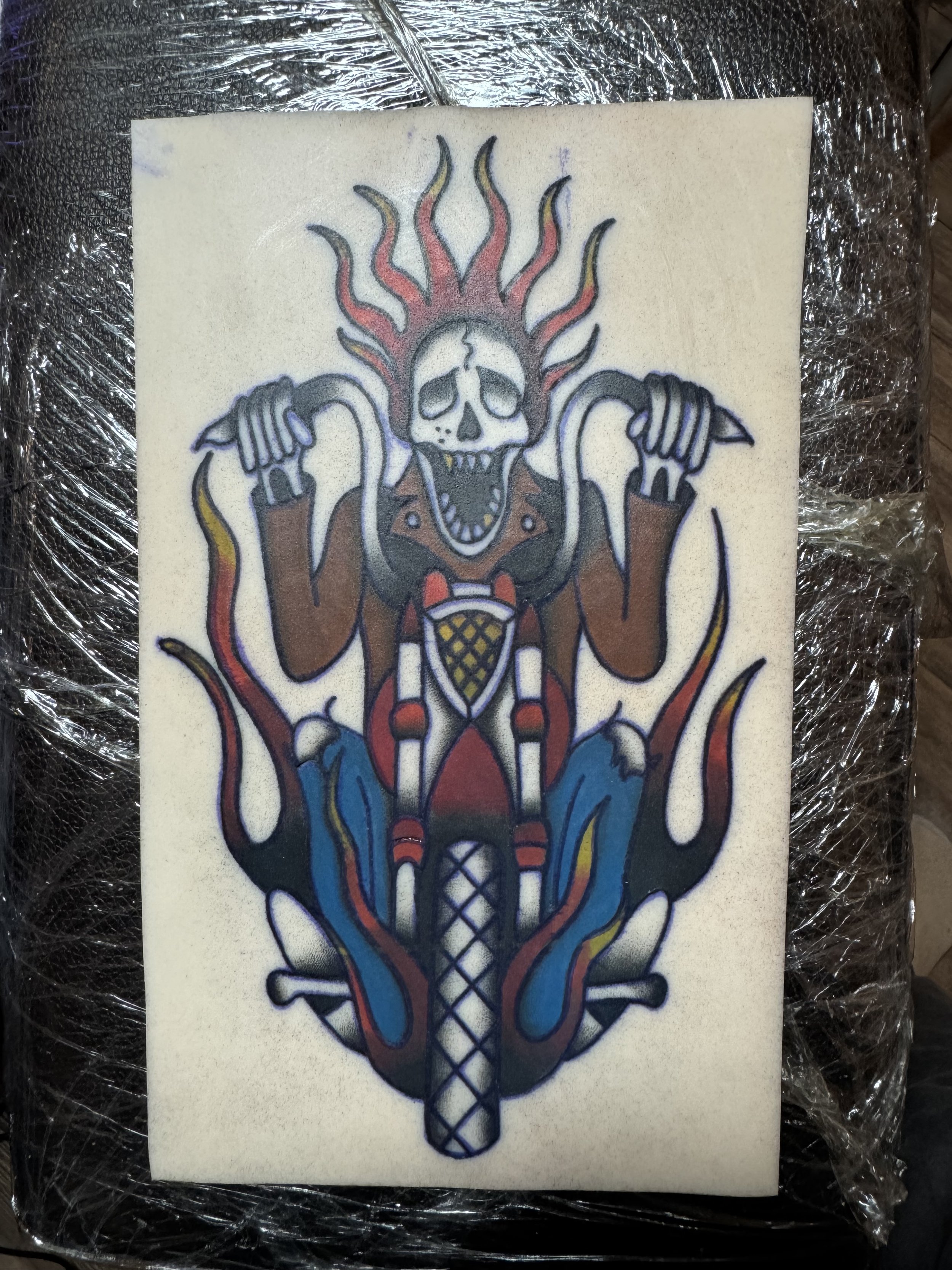 A tattoo design featuring a skeleton with a flaming skull riding a motorcycle, with flames and smoke details.