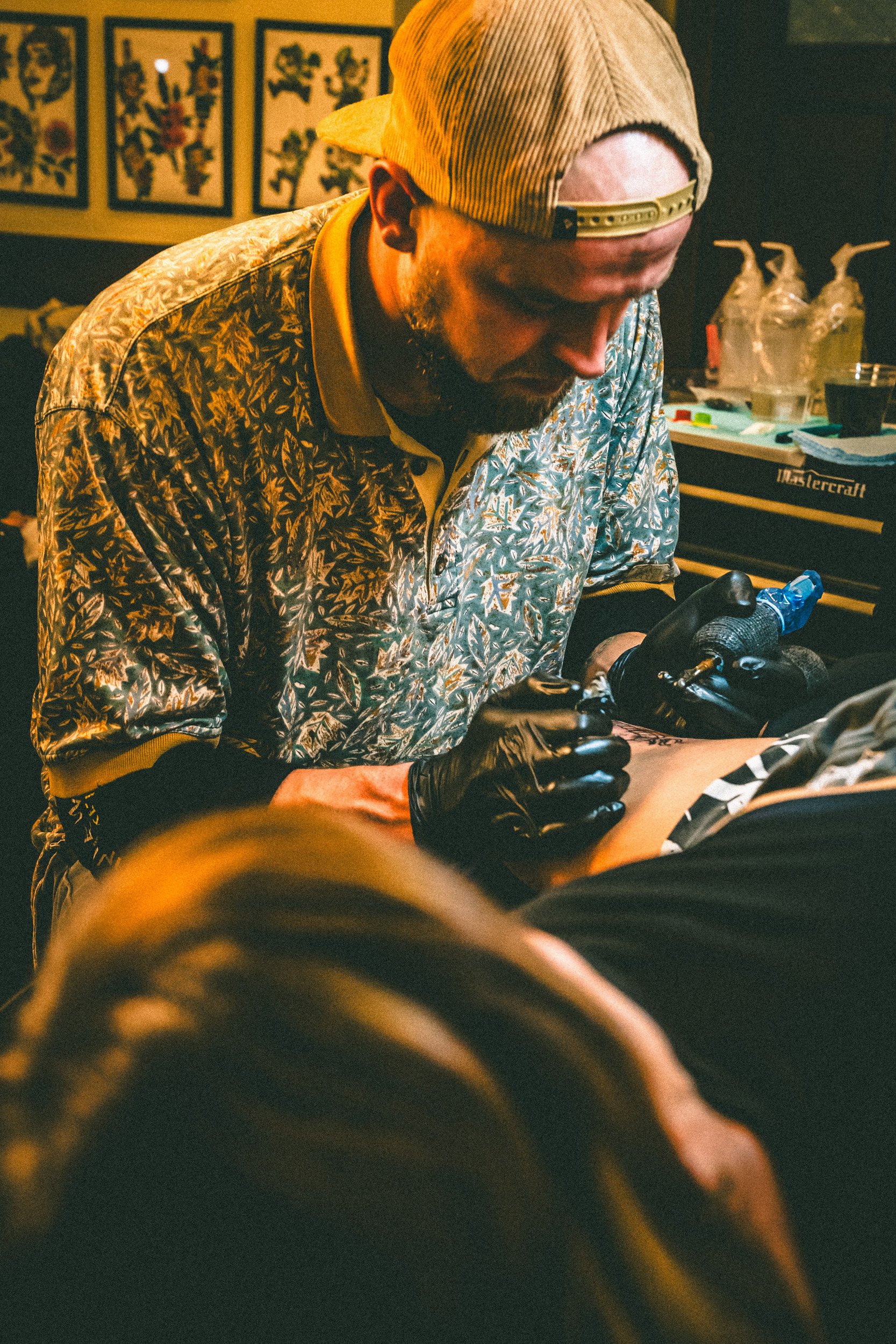 A man with a tattoo sleeve over his shirt, wearing a backwards baseball cap and black gloves, is tattooing a person's arm. The background shows framed pictures on the wall and tattoo supplies on a table.