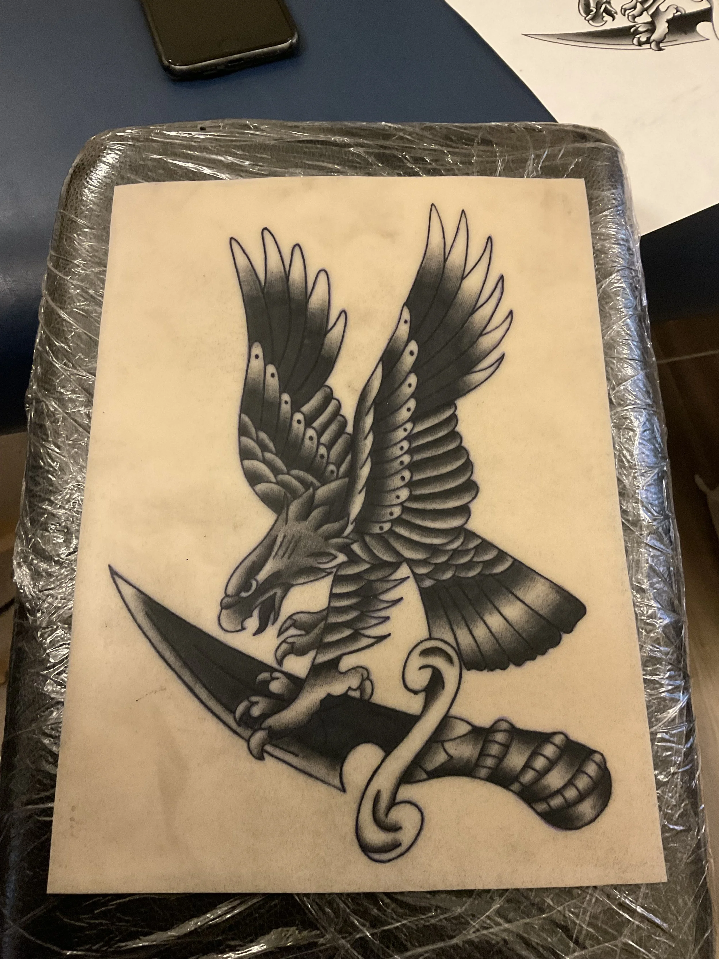 A black and gray tattoo design of a fierce eagle with wings spread, grasping a sword in its talons, with a snake curled around the sword.