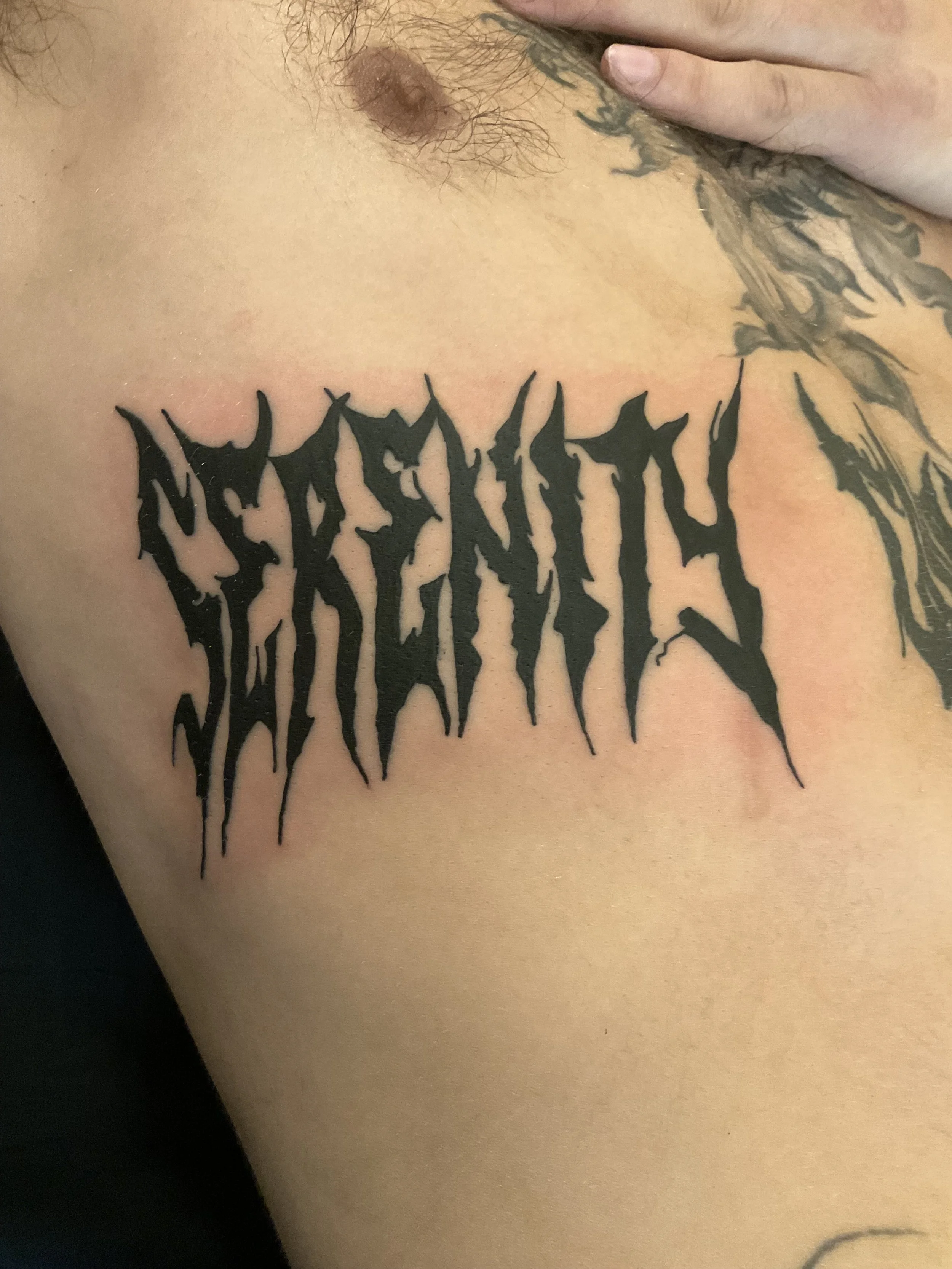 Tattoo reading "RESIST" in black Gothic-style font on a person's chest, with part of a tattooed arm and additional tattoos visible.