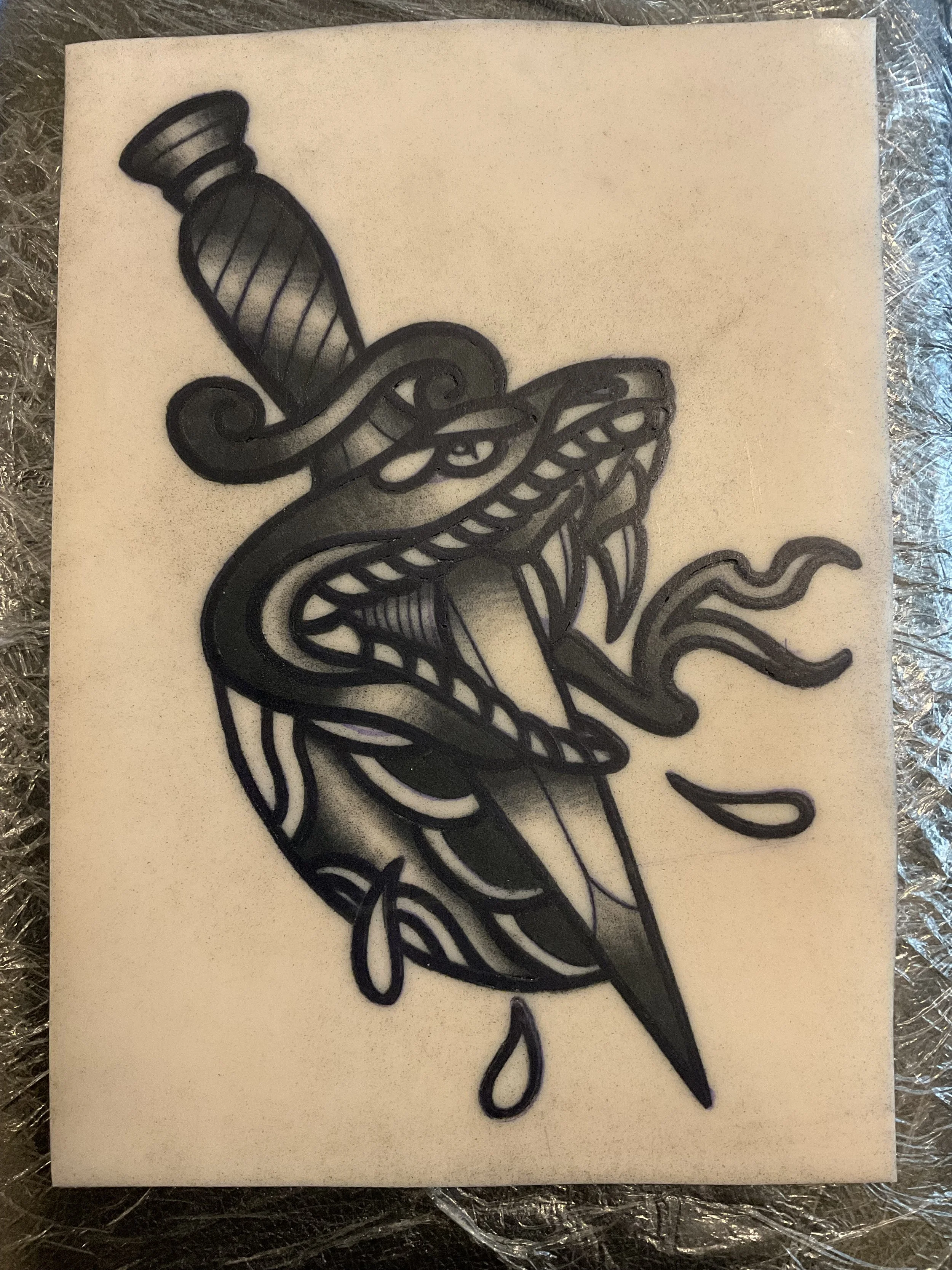 Black and gray tattoo design of a snake wrapped around a dagger on a piece of white paper.