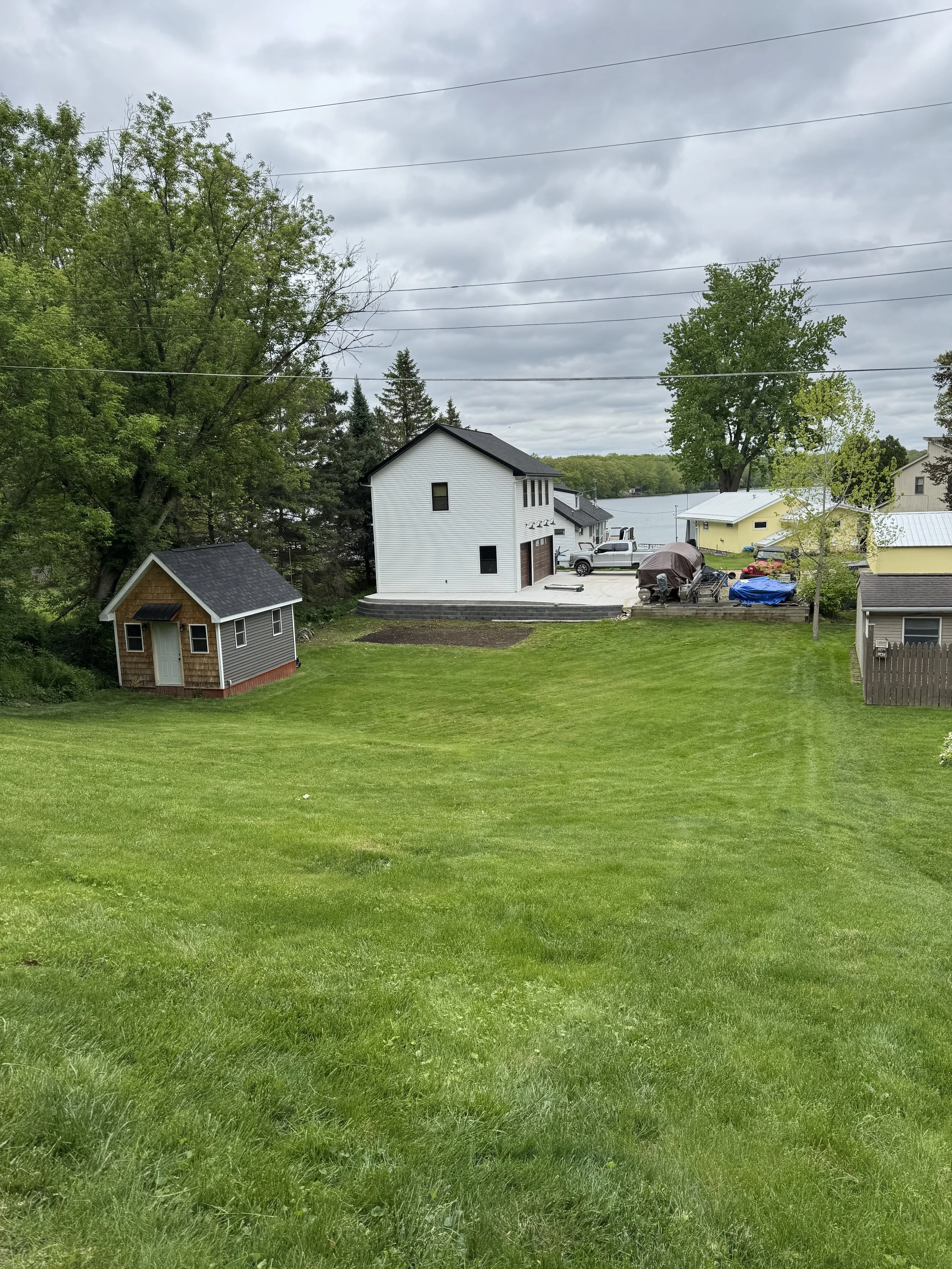A backyard with well-maintained green grass, a small shed to the left, and a white house with a driveway leading to a waterfront in the background. Trees and cloudy sky are visible.