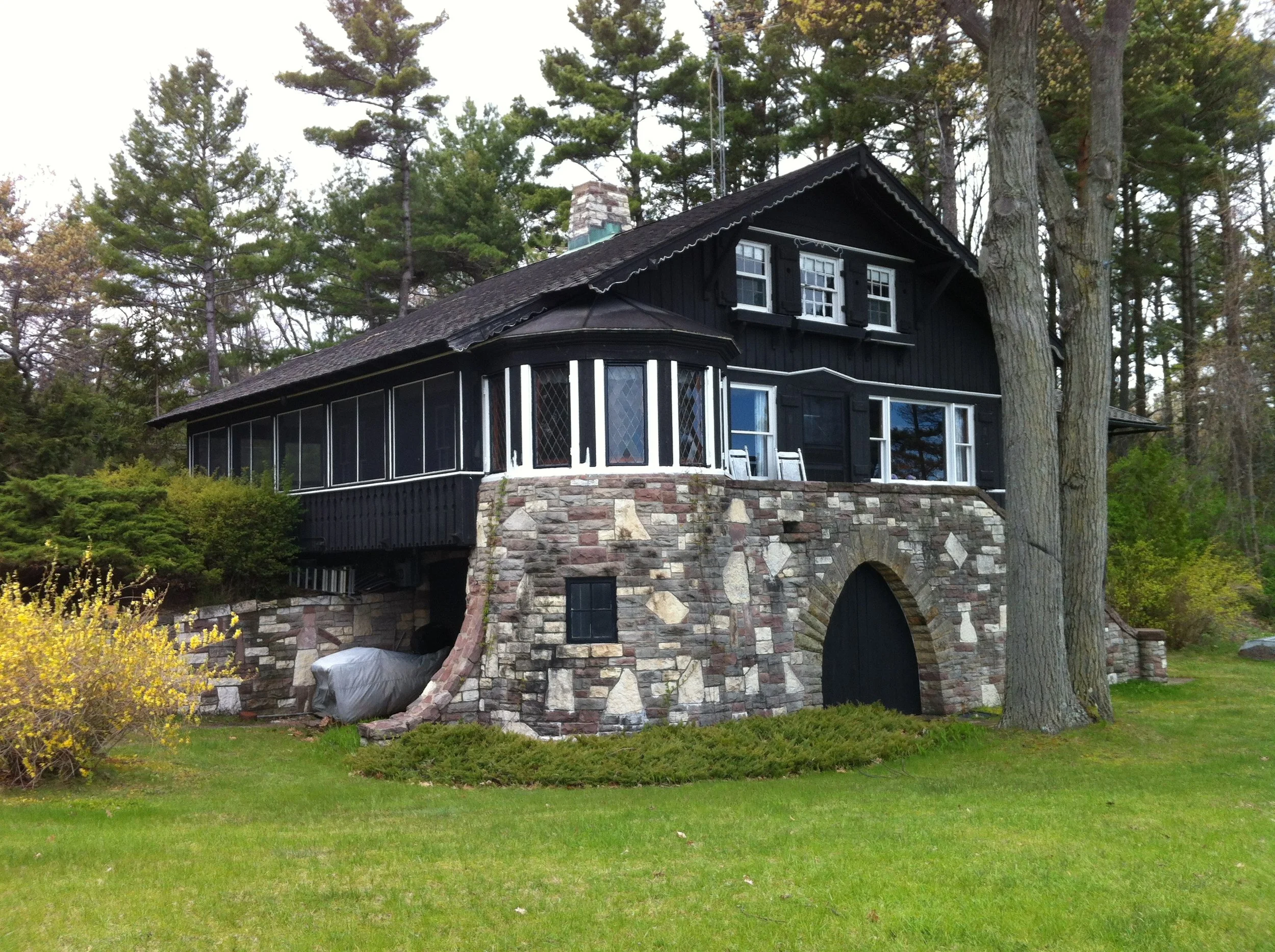 A large house with black wooden siding and a stone foundation, featuring a rounded porch with screens, surrounded by trees and a well-maintained lawn.