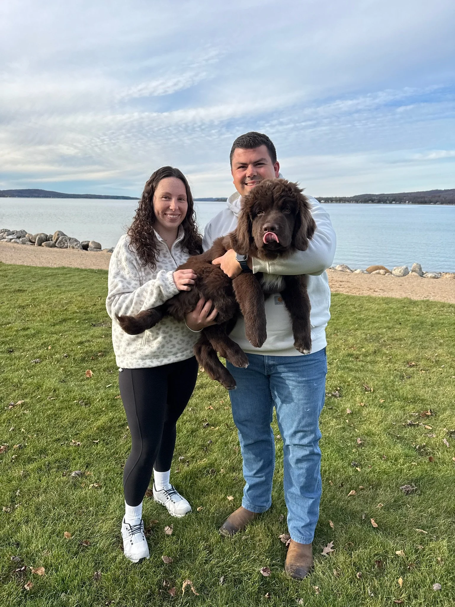 A happy couple outdoors by a lake, holding a large brown Newfoundland puppy, with a partly cloudy sky in the background.