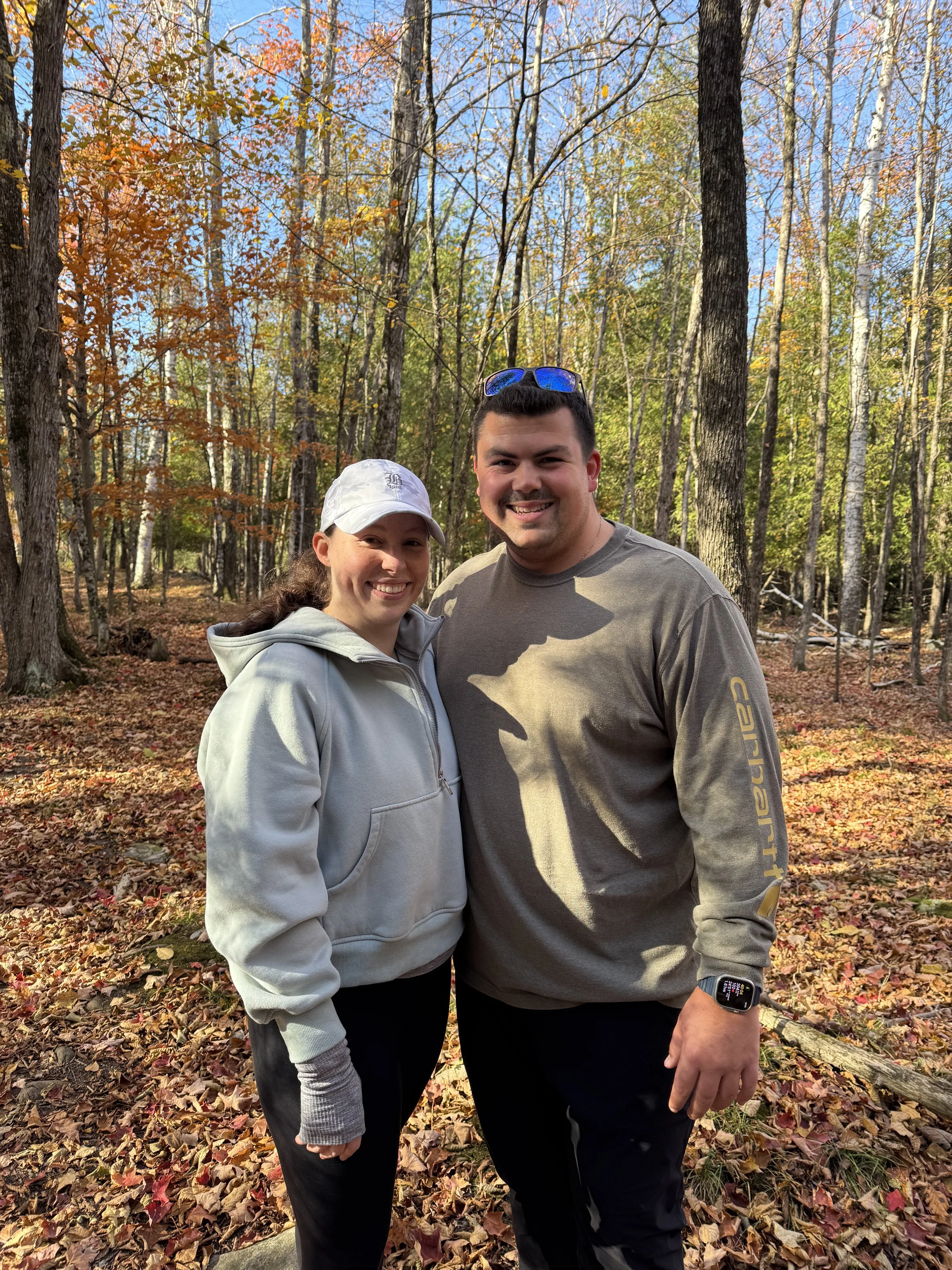 A smiling man and woman standing in a fall wooded forest, surrounded by colorful leaves and trees, with blue sky above.