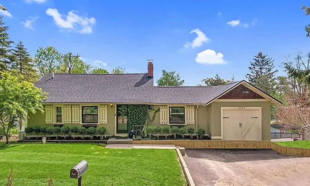 A single-story house with a green exterior, dark gray roof, and a white garage door. The front yard features a well-maintained lawn, some bushes, and a small tree, with a pathway leading to the main entrance and driveway on the right side. The background includes tall trees and a partly cloudy blue sky.