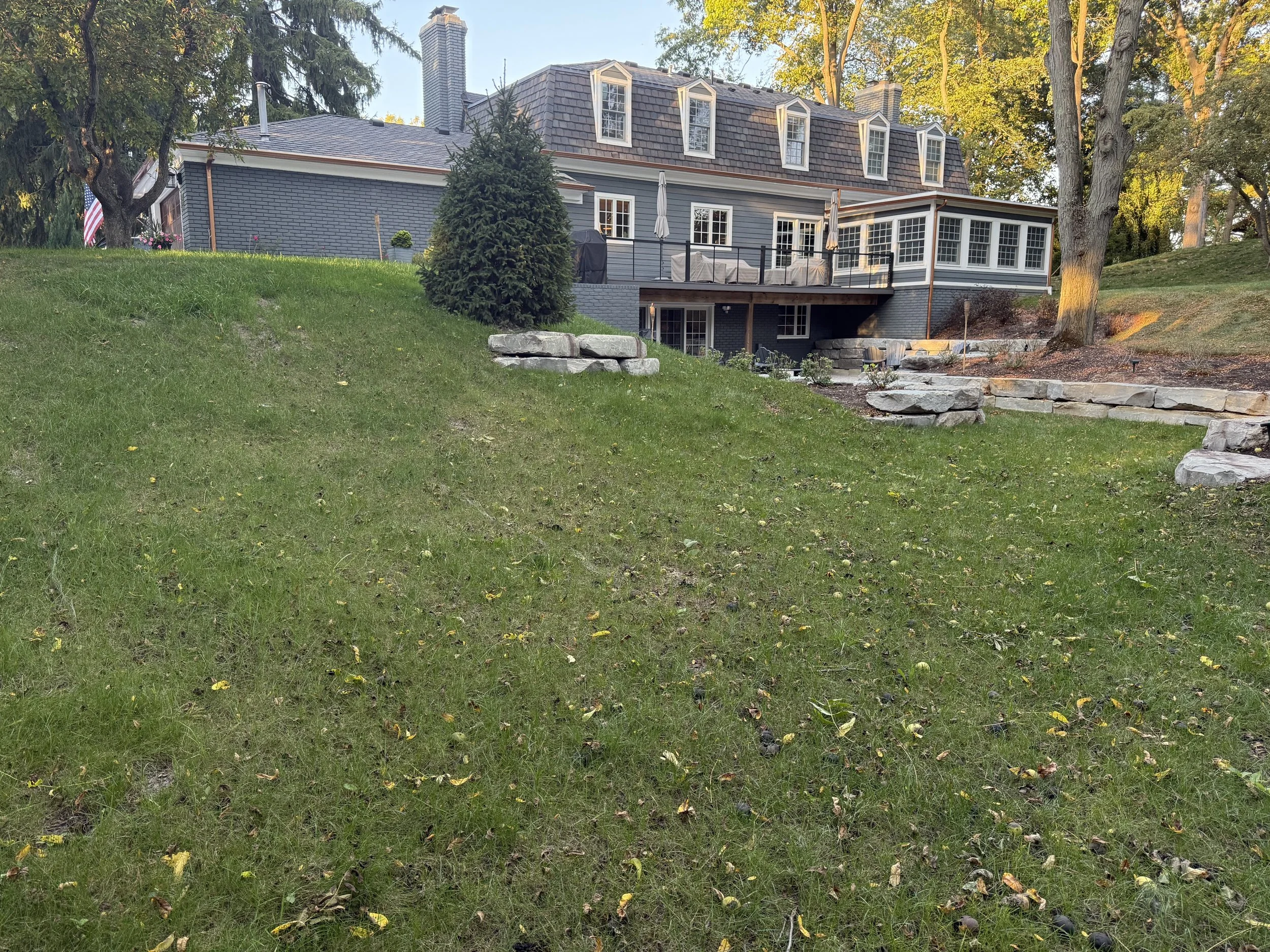 Backyard with a sloped grassy lawn, stone retaining walls, large trees, and a blue house with a deck and multiple windows.